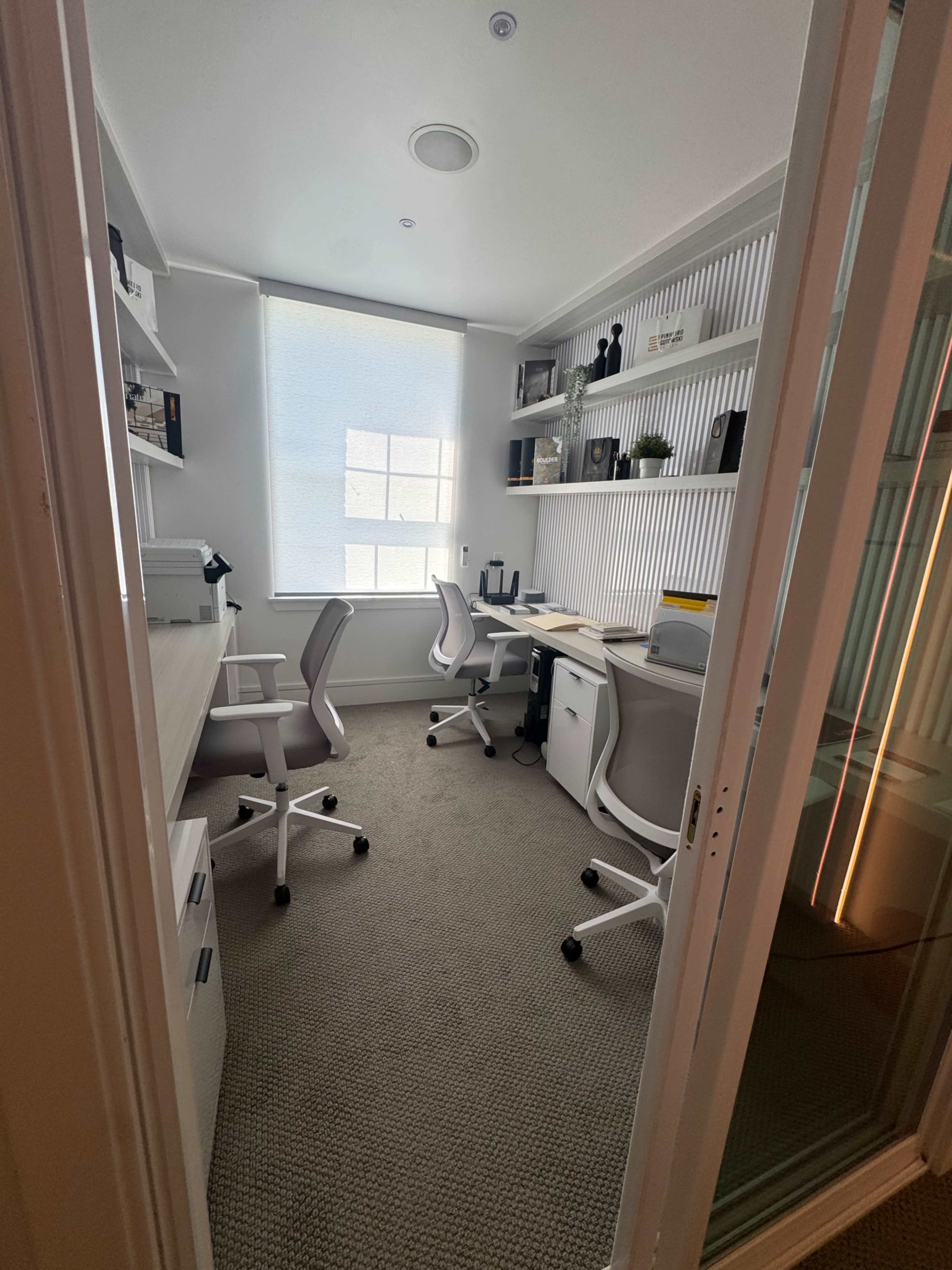 The image shows a small home office with two desks, ergonomic chairs, shelves, and a window providing natural light.
