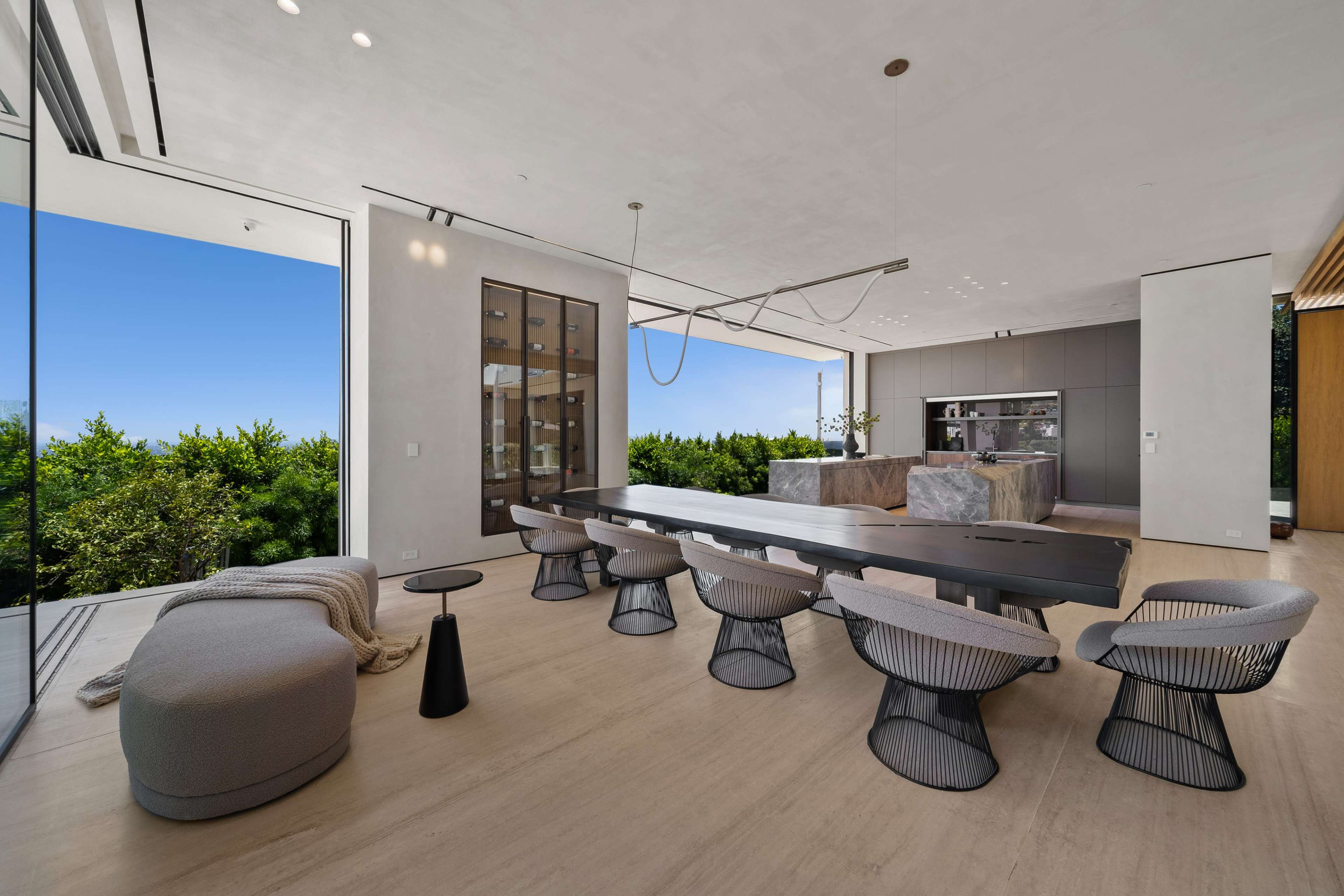 The image shows a modern dining area with a long table, surrounded by unique chairs, and large windows that provide a view of greenery outside.