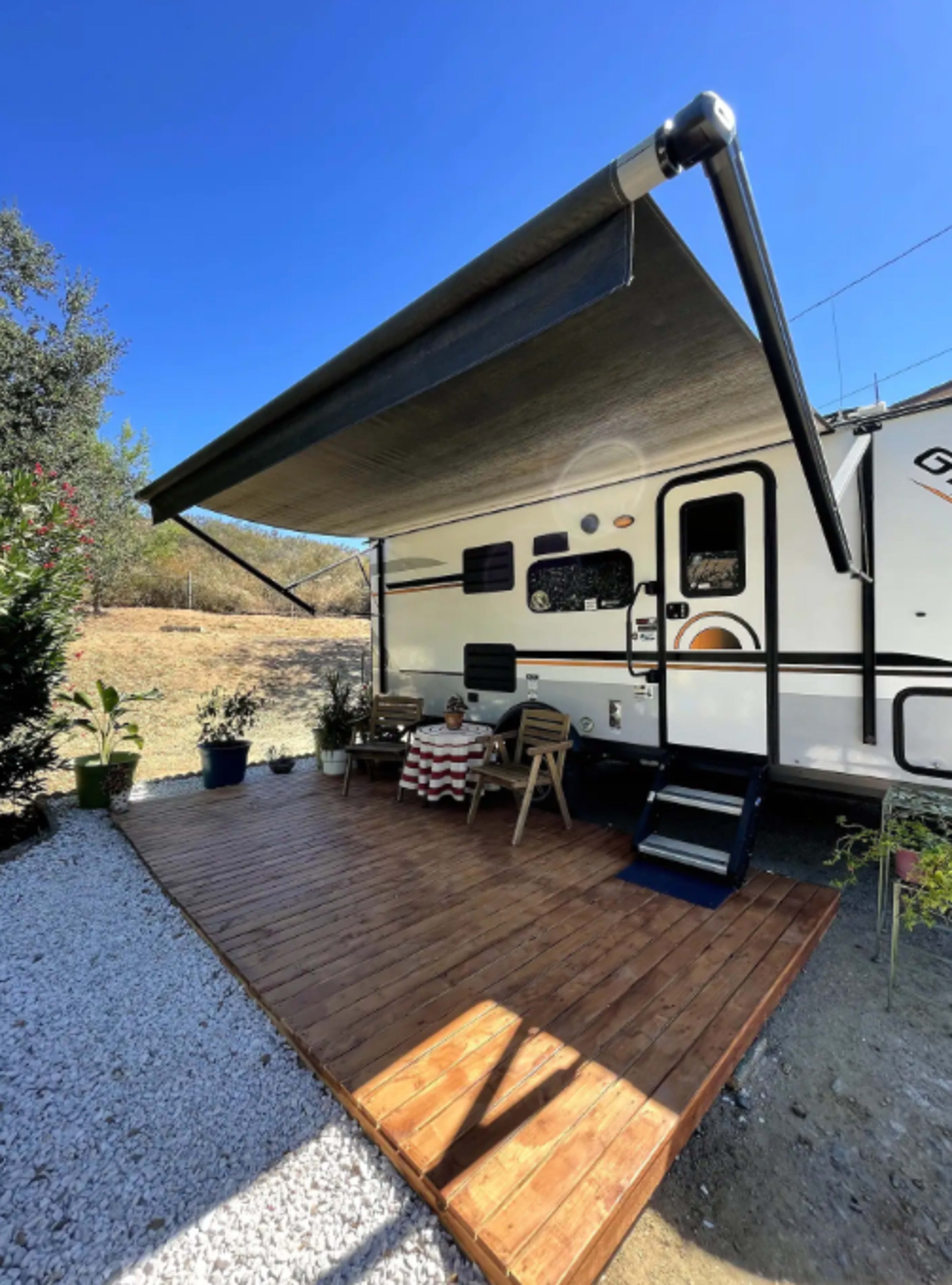 A recreational vehicle with an extended awning sits on a wooden deck surrounded by potted plants and gravel.