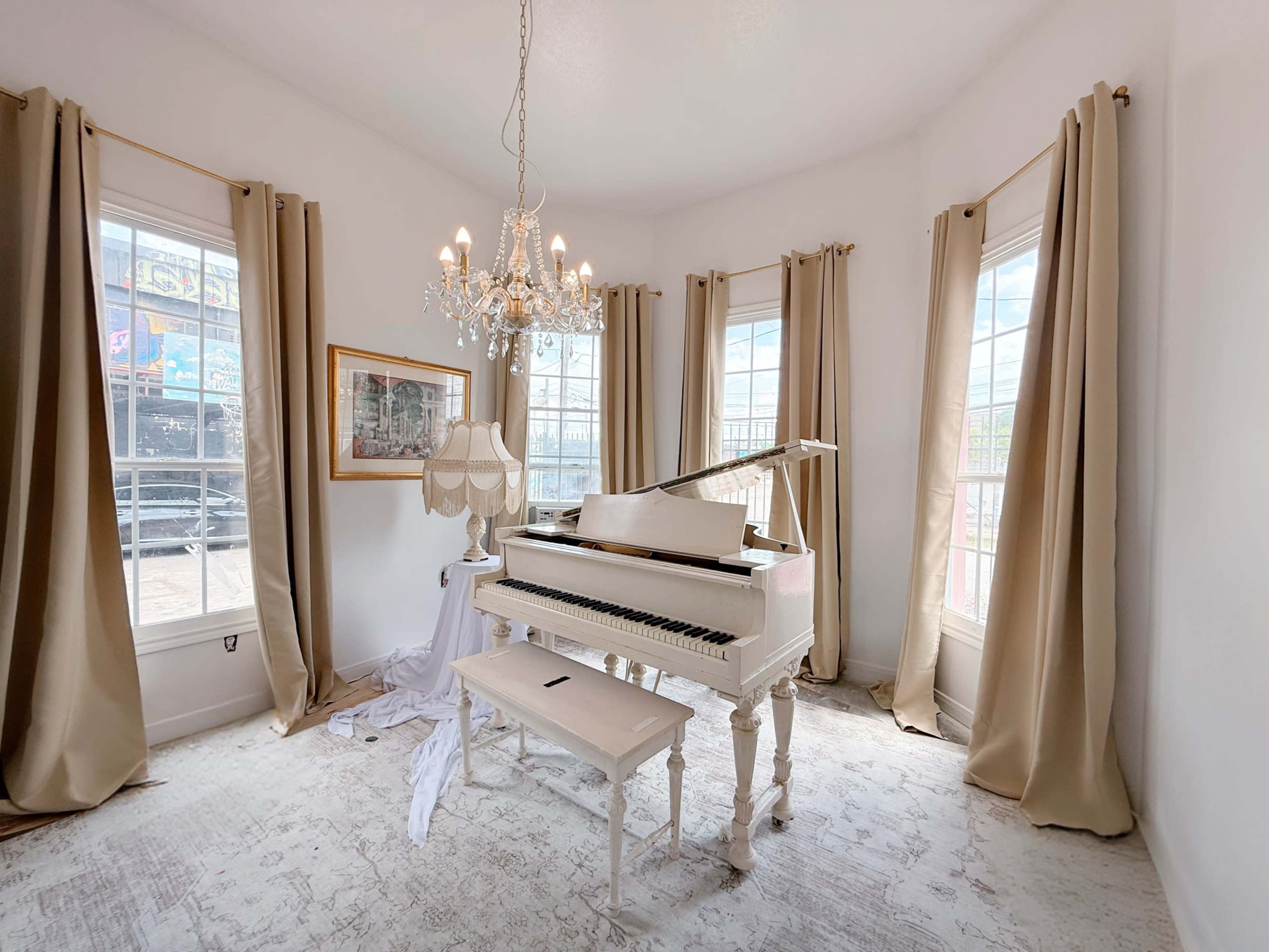 A white grand piano is situated in a bright room with large windows, surrounded by beige curtains and a chandelier.