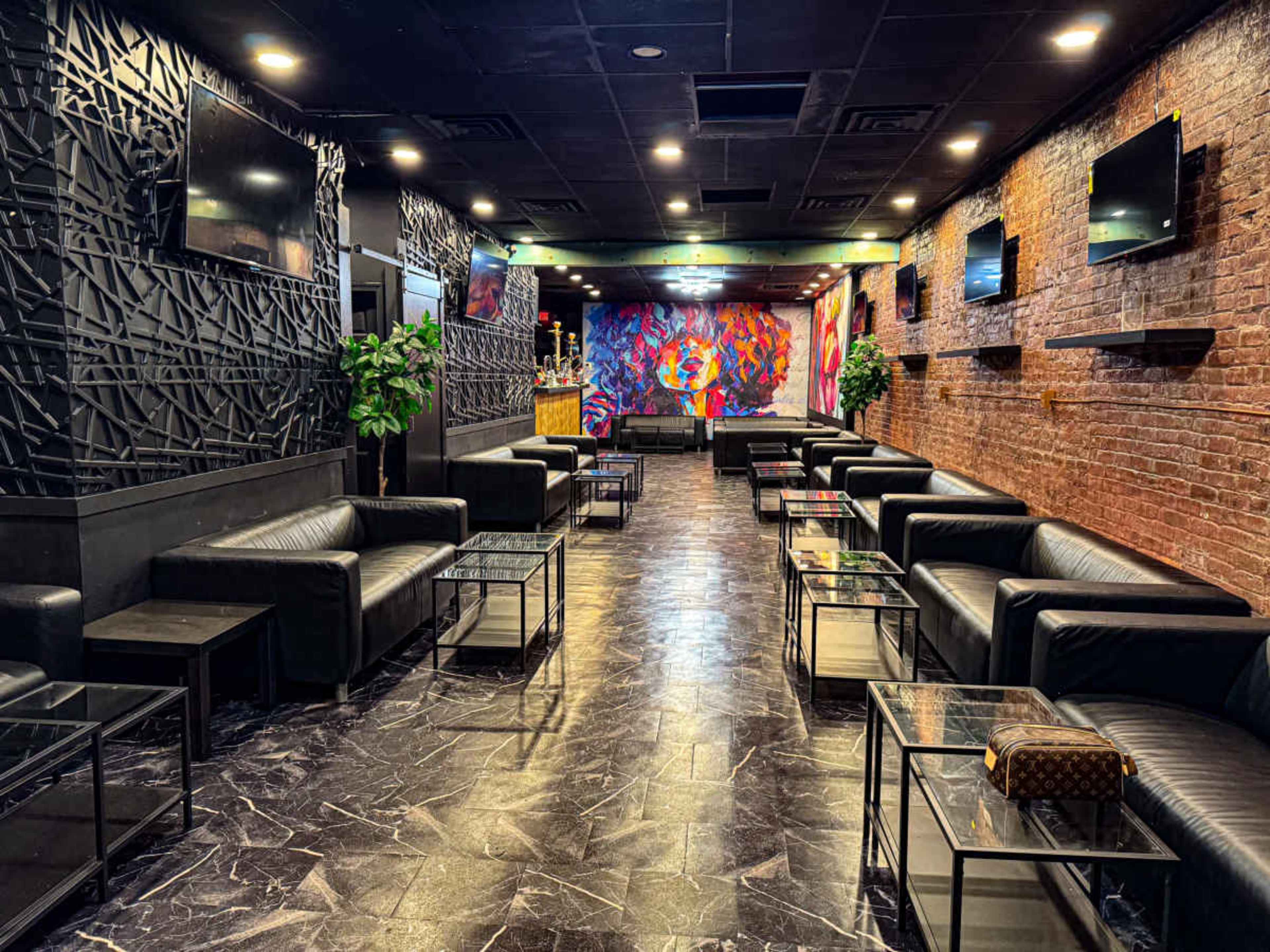 The image shows a modern lounge space with black leather seating, glass tables, and vibrant wall art, illuminated by overhead lights.
