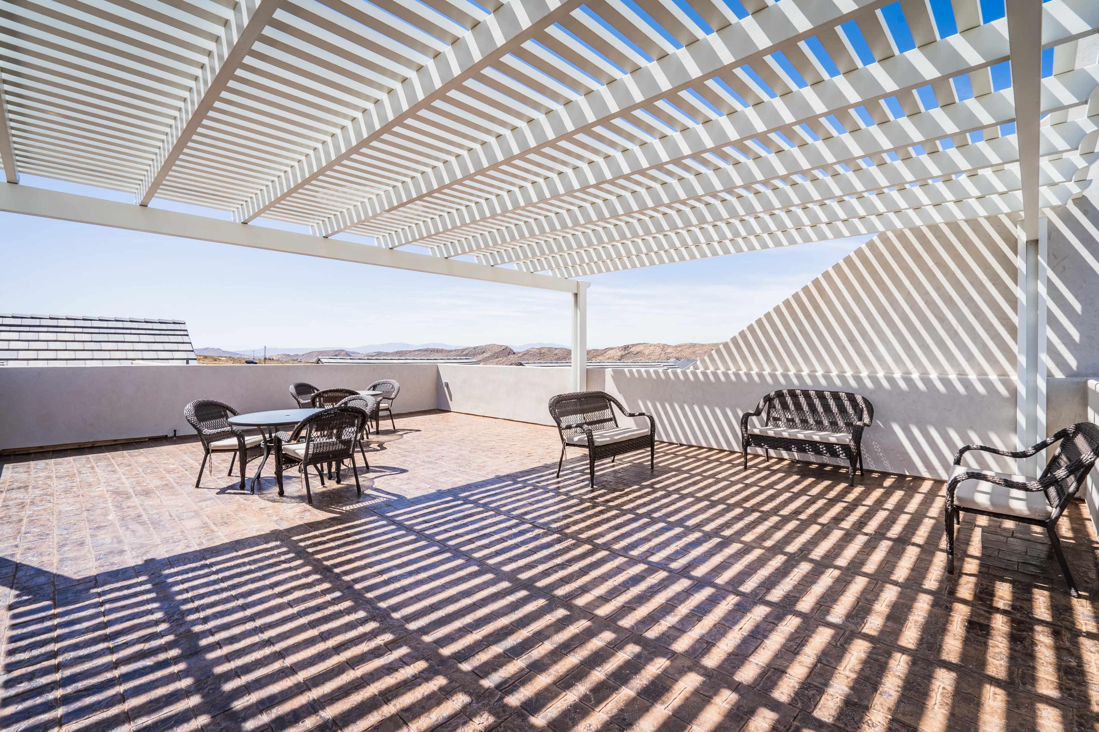 A spacious rooftop terrace features dining and seating areas beneath a slatted pergola, with sunlight creating striped shadows on the tiled floor.