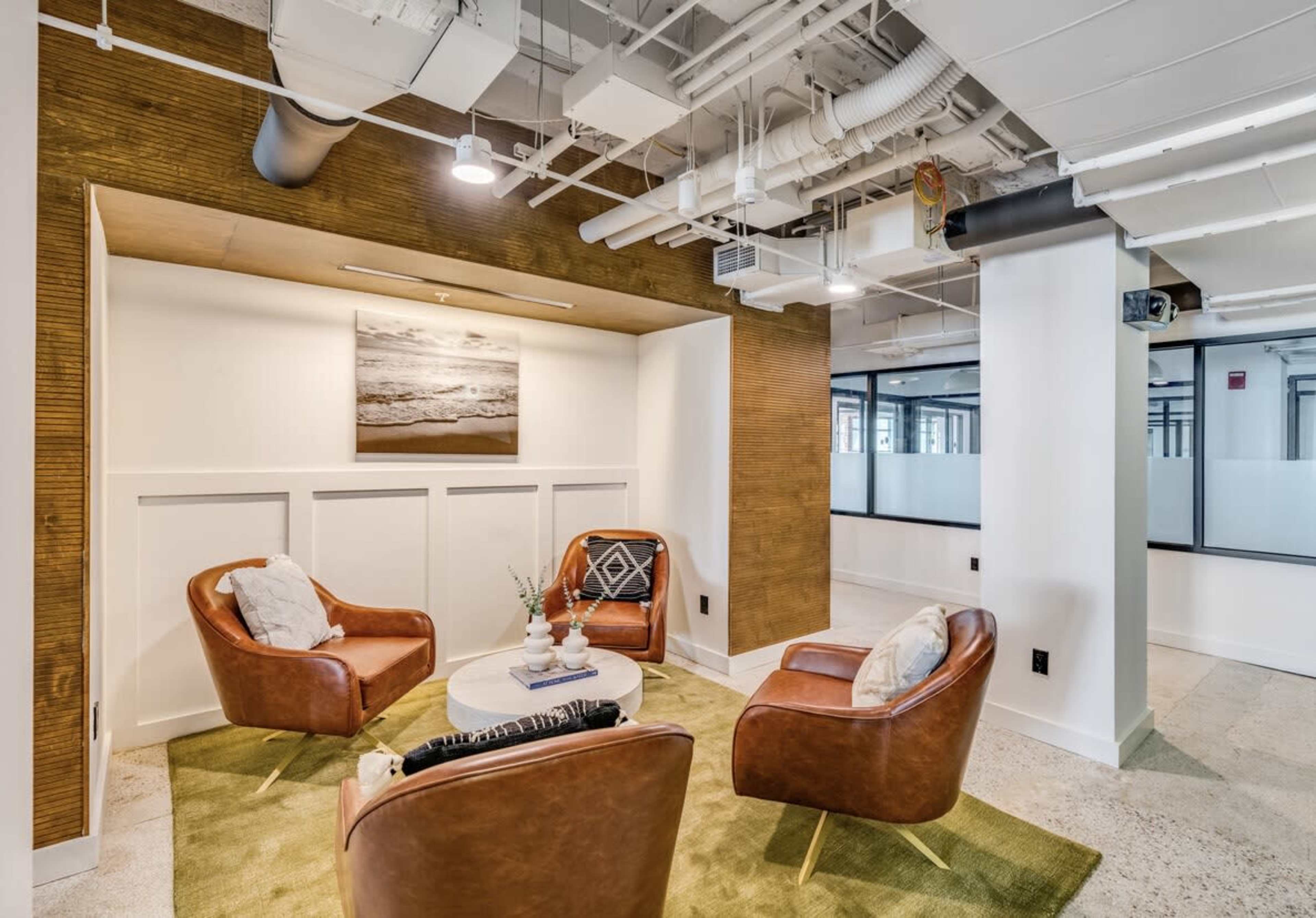 A modern office lounge area with three brown leather chairs arranged around a small circular table on a green rug, featuring a wall art piece and a minimalistic decor.