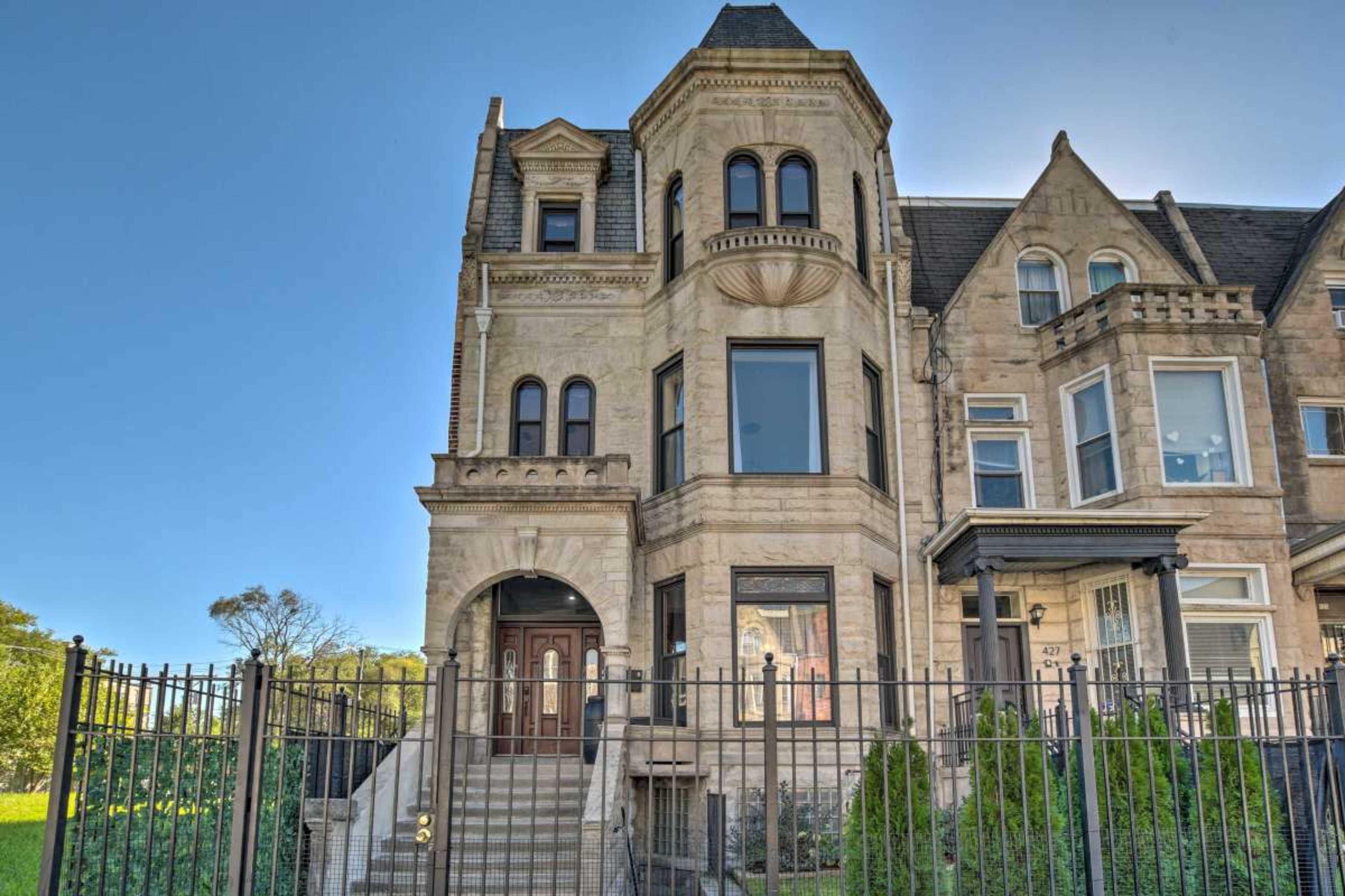 The image shows a large, ornate stone house with a pointed roof and a decorative facade, situated behind a wrought iron fence.