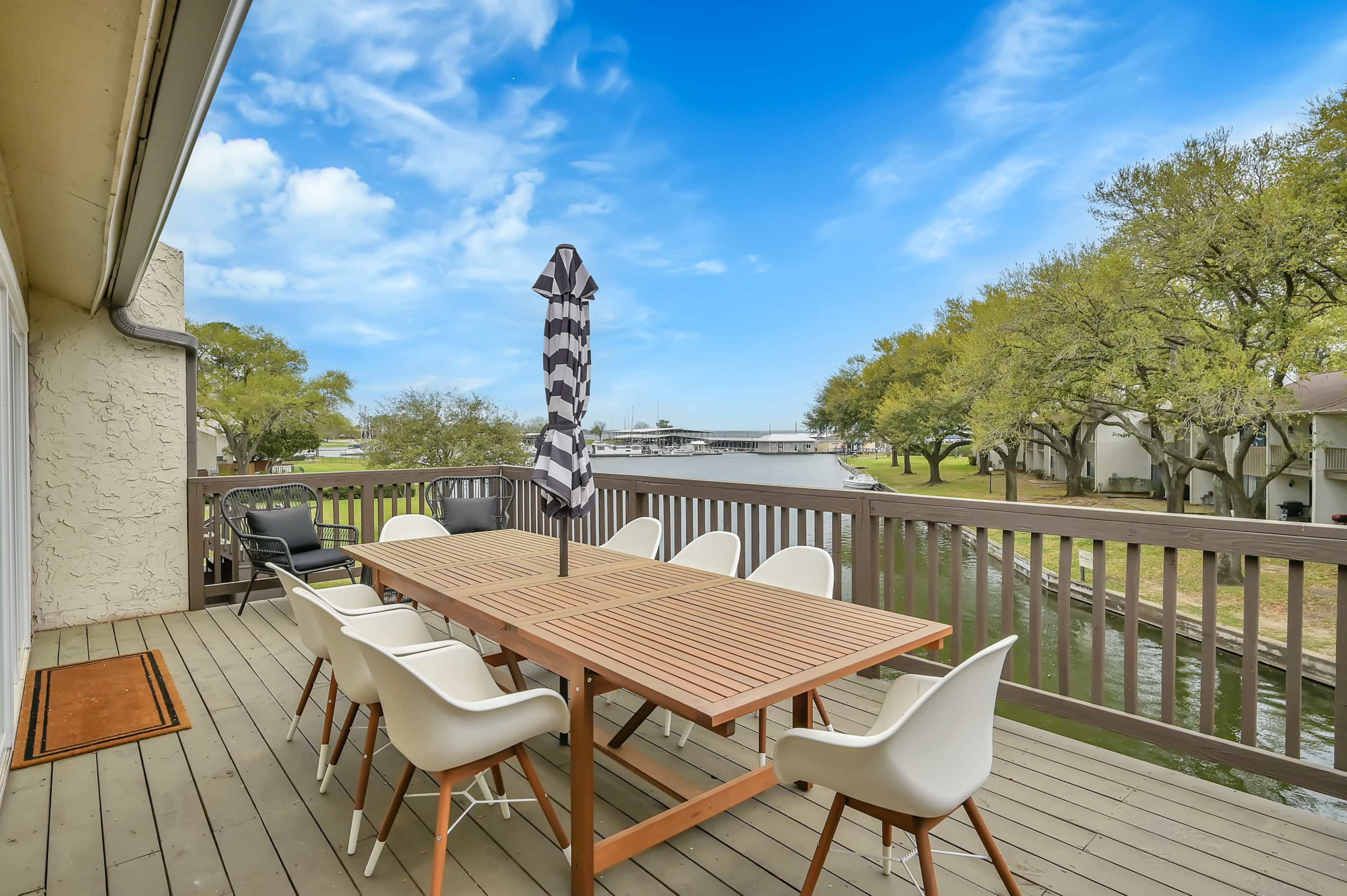 A wooden deck features a large dining table surrounded by white chairs, overlooking a waterway lined with trees.