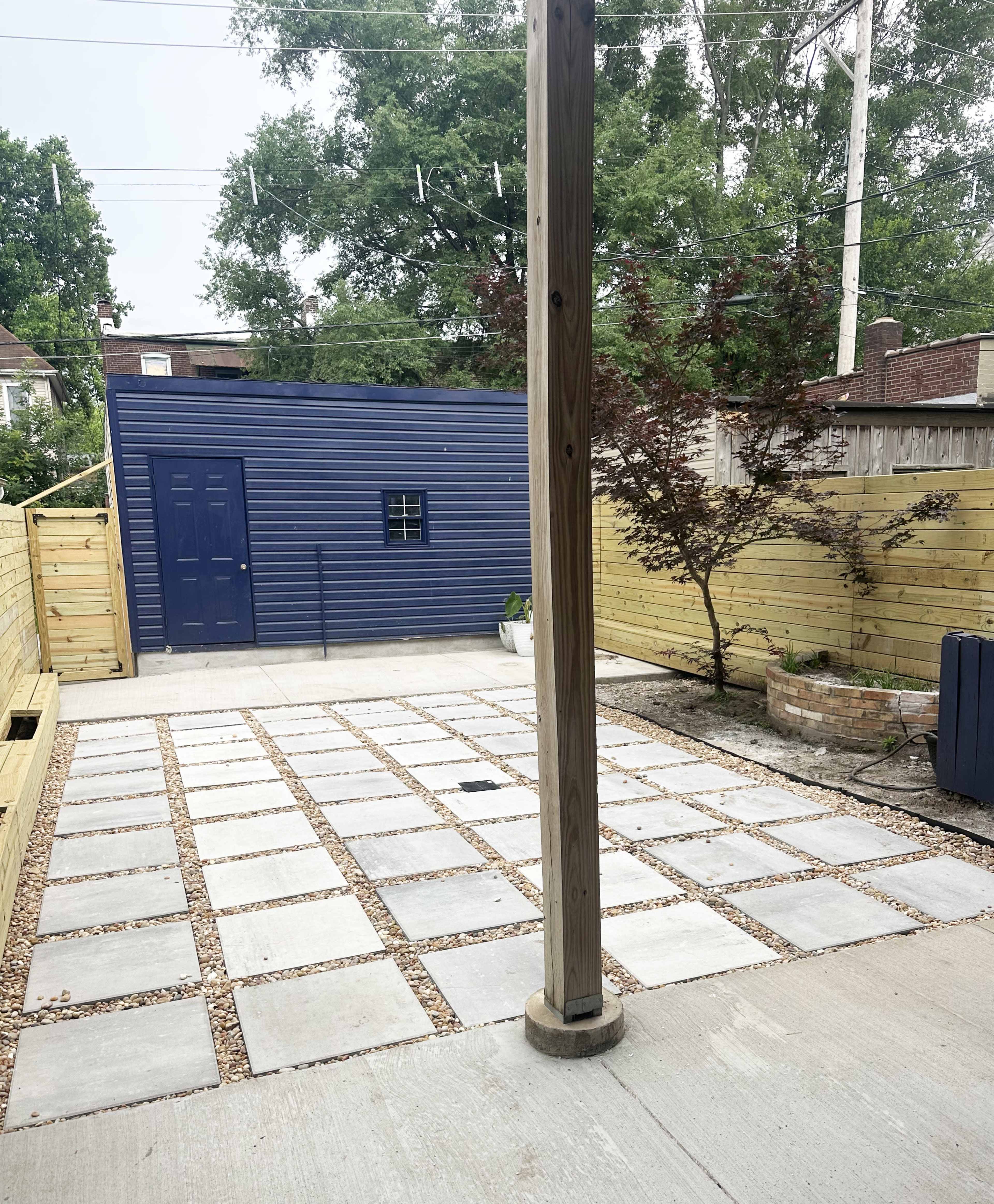 The image shows a modern outdoor patio area with a gravel pathway bordered by large concrete pavers, leading to a blue shed in the background, surrounded by wooden fencing and greenery.