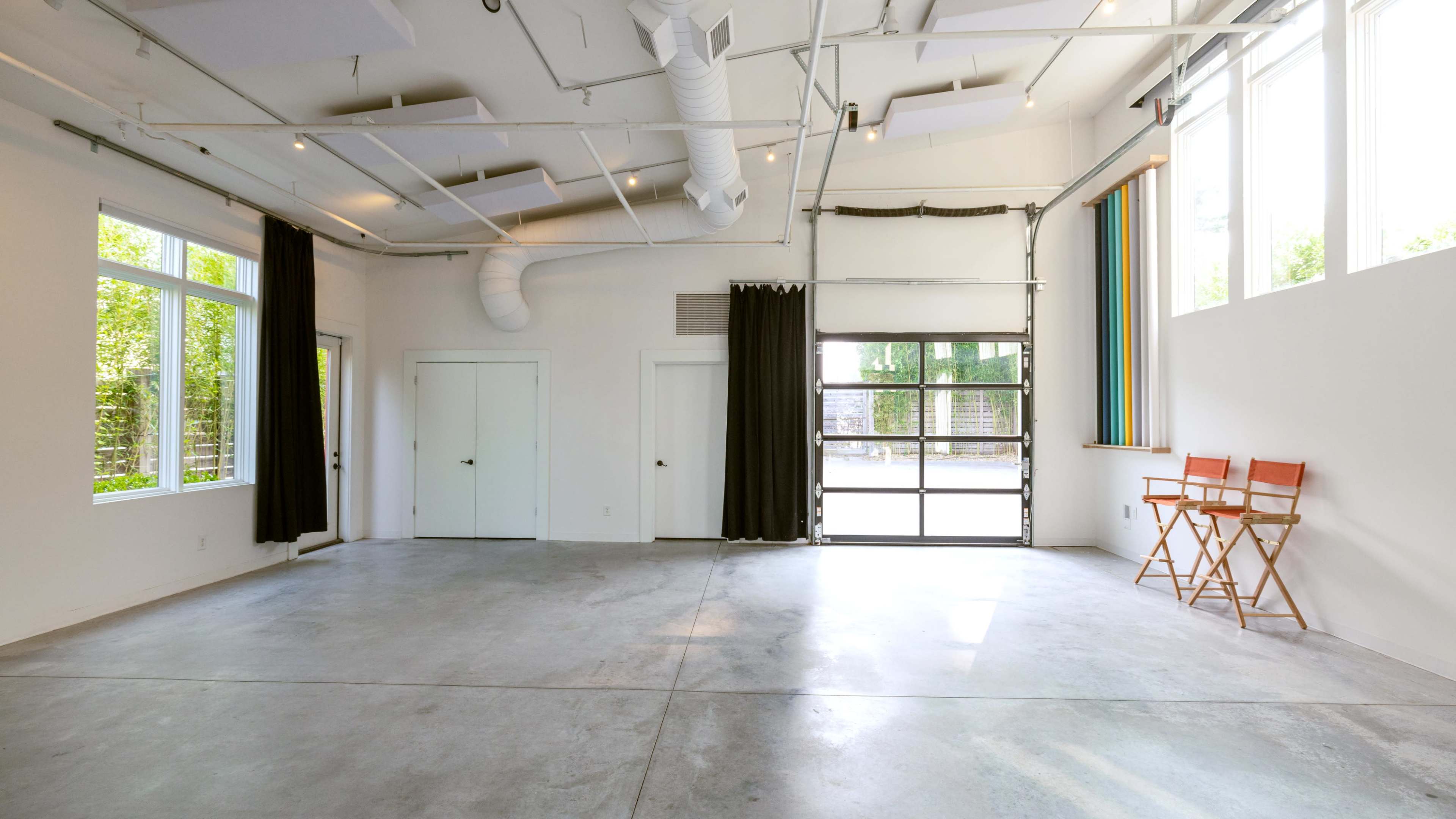 The image shows an empty, modern garage space with large windows, concrete flooring, and two directors' chairs positioned beside an open garage door.