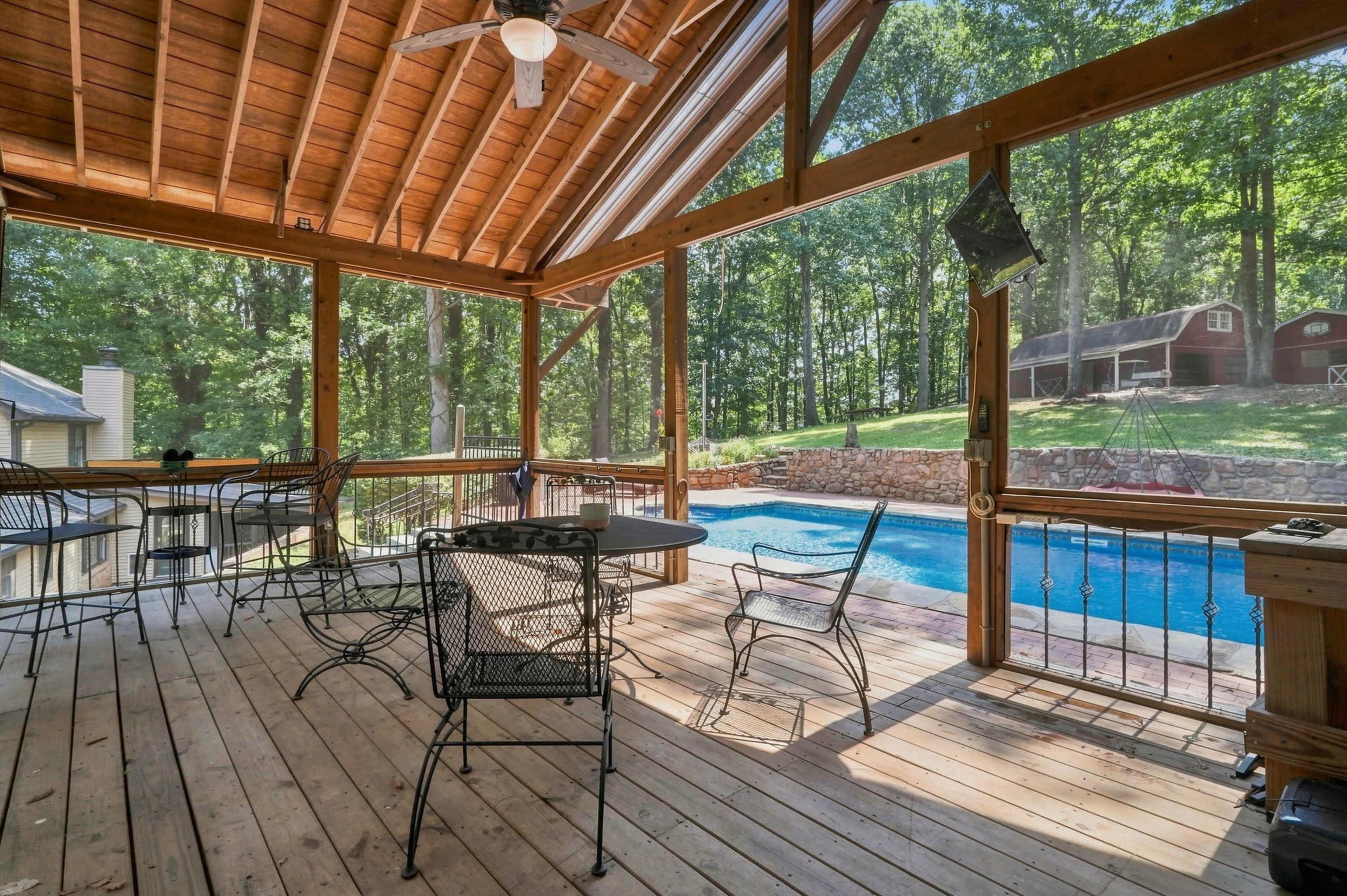 A screened porch with wooden flooring and a ceiling fan overlooks a pool and a wooded area in the background.