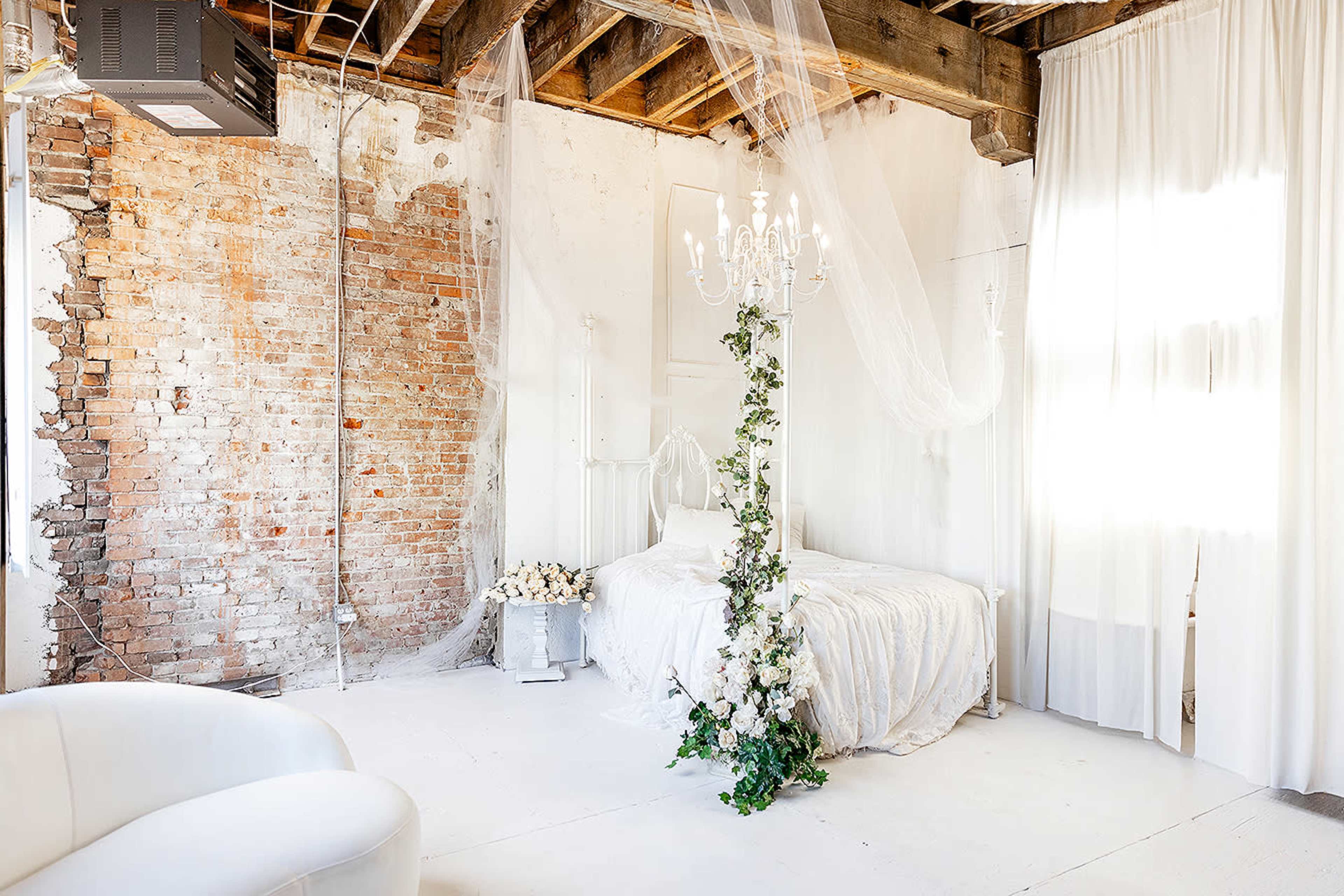 A bright bedroom with exposed brick walls, a white bed adorned with floral decorations, and sheer drapes hanging from the ceiling.