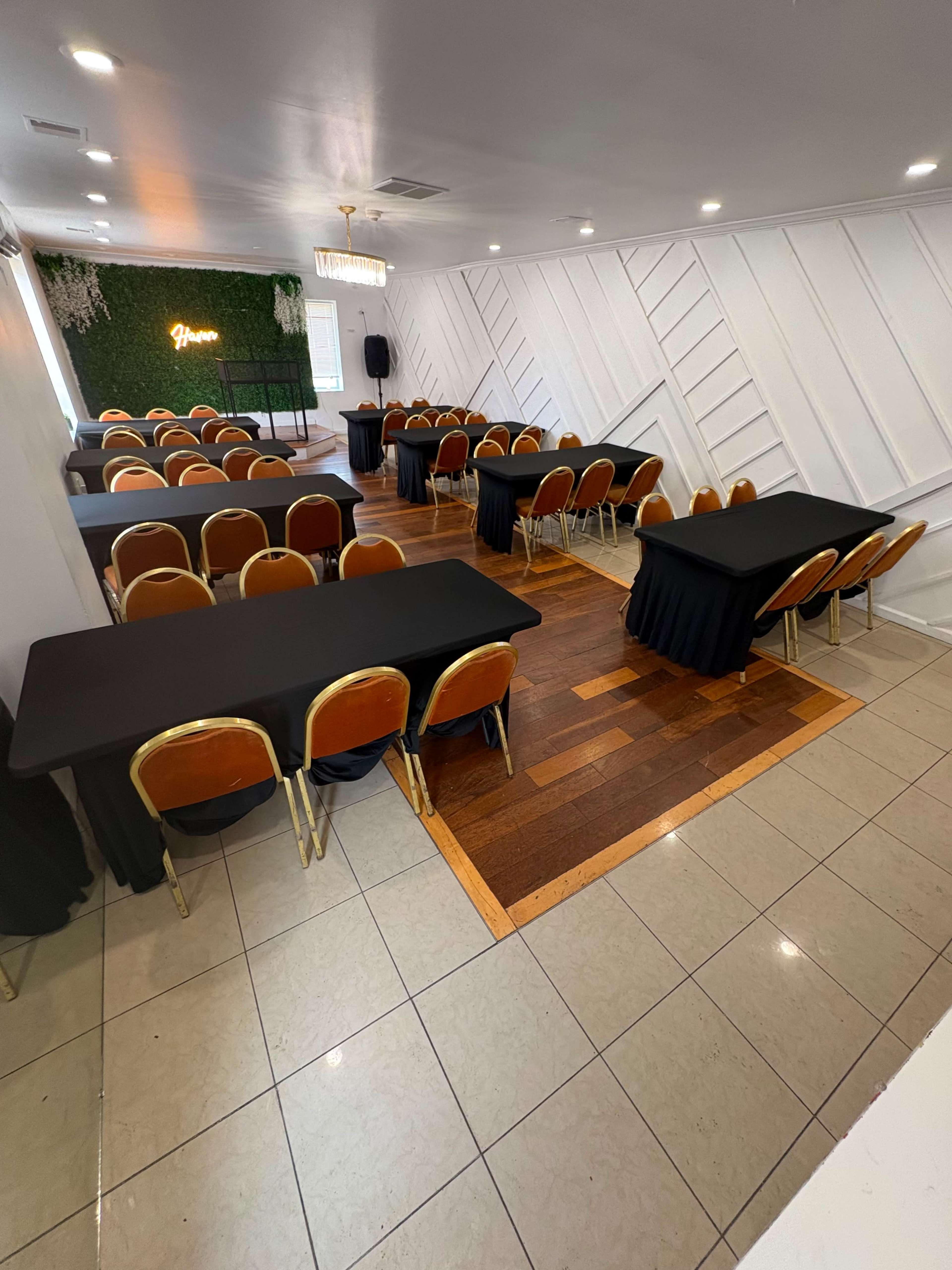 The image shows a spacious event room set up with several tables covered in black tablecloths and chairs arranged in a semi-circle, with a decorative green wall and a chandelier in the background.