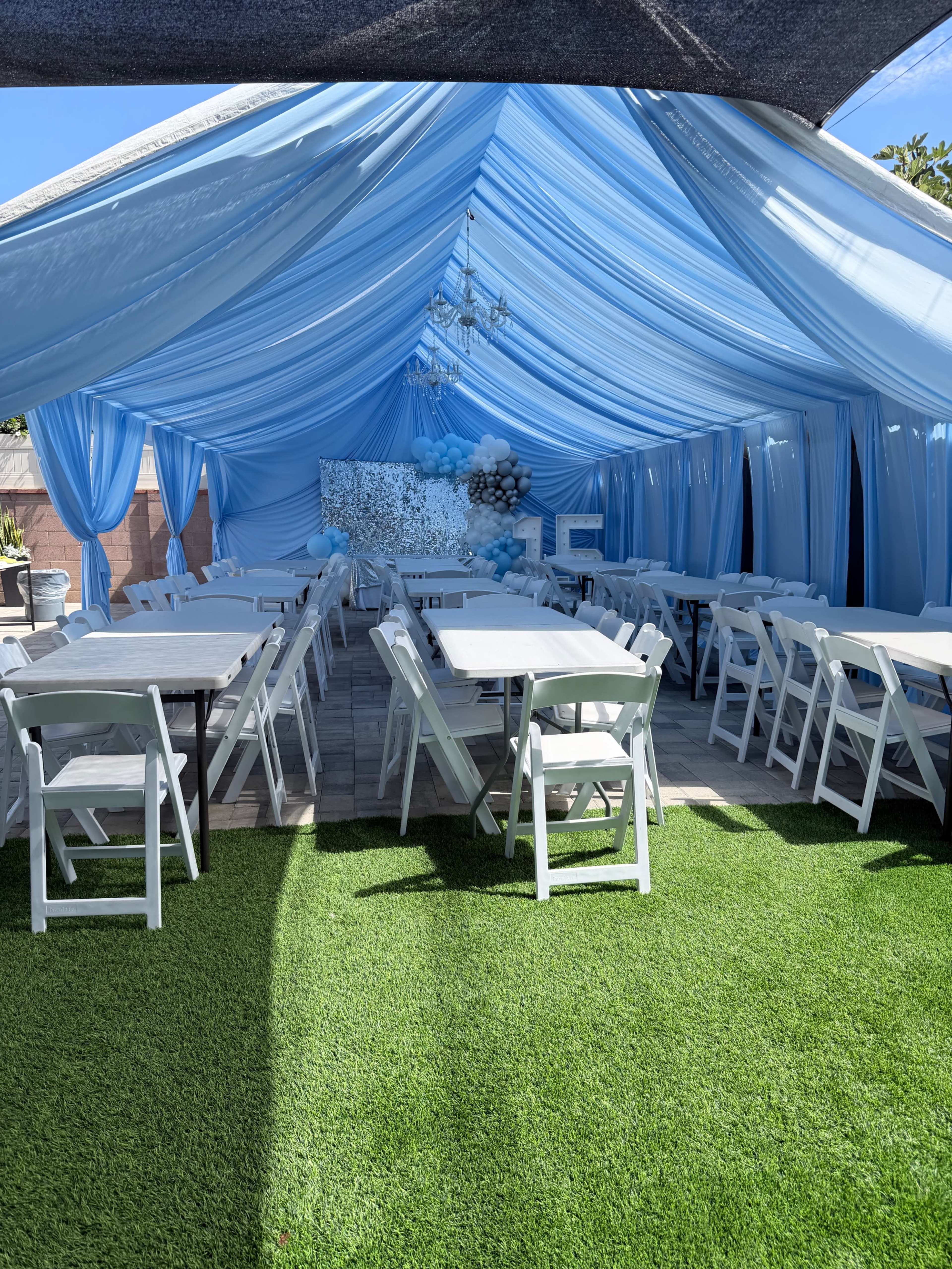 The image shows a tented outdoor space decorated in blue and white, featuring white tables and folding chairs arranged on a grass-like surface.