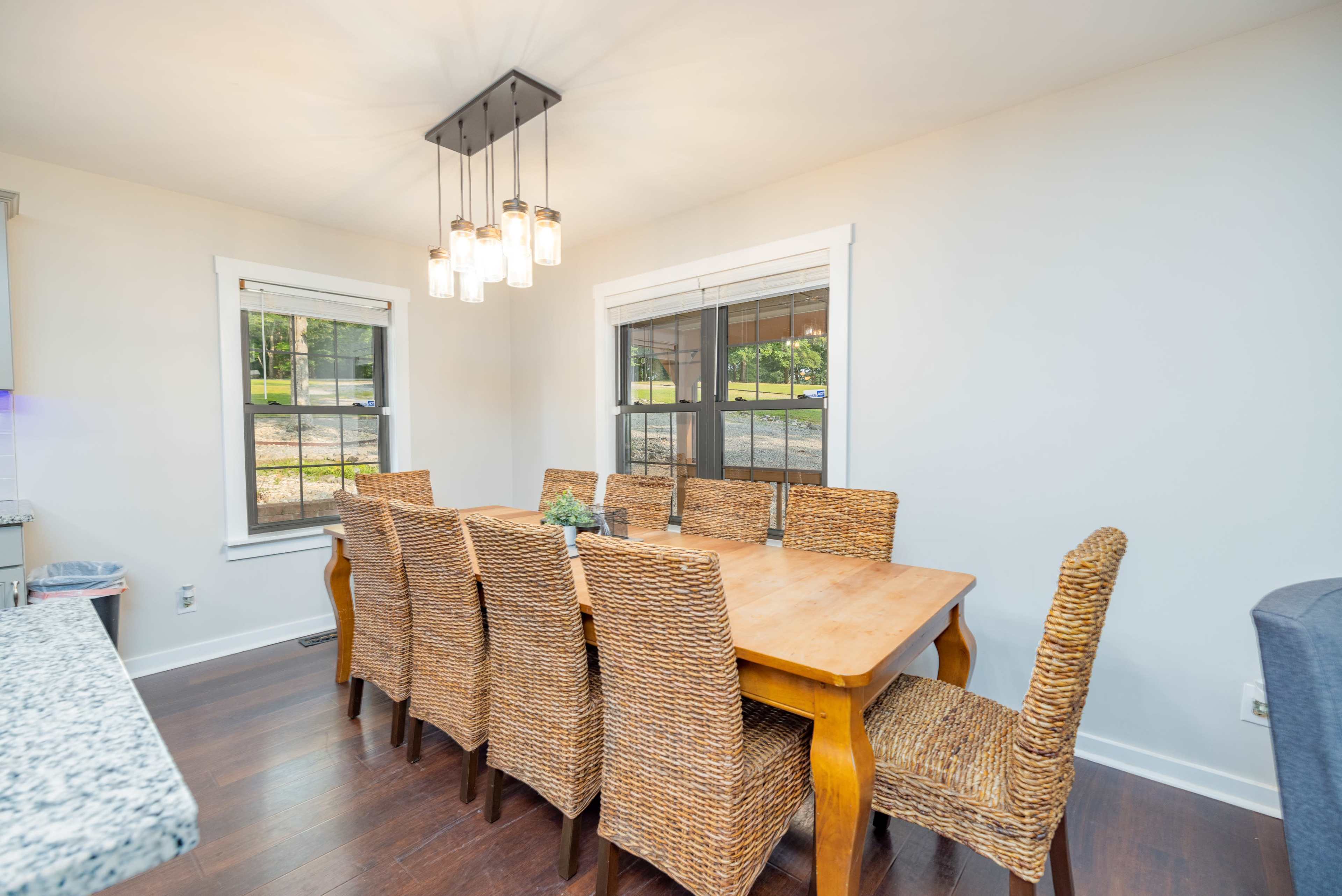 The image shows a dining area with a large wooden table surrounded by woven chairs and illuminated by pendant lighting.