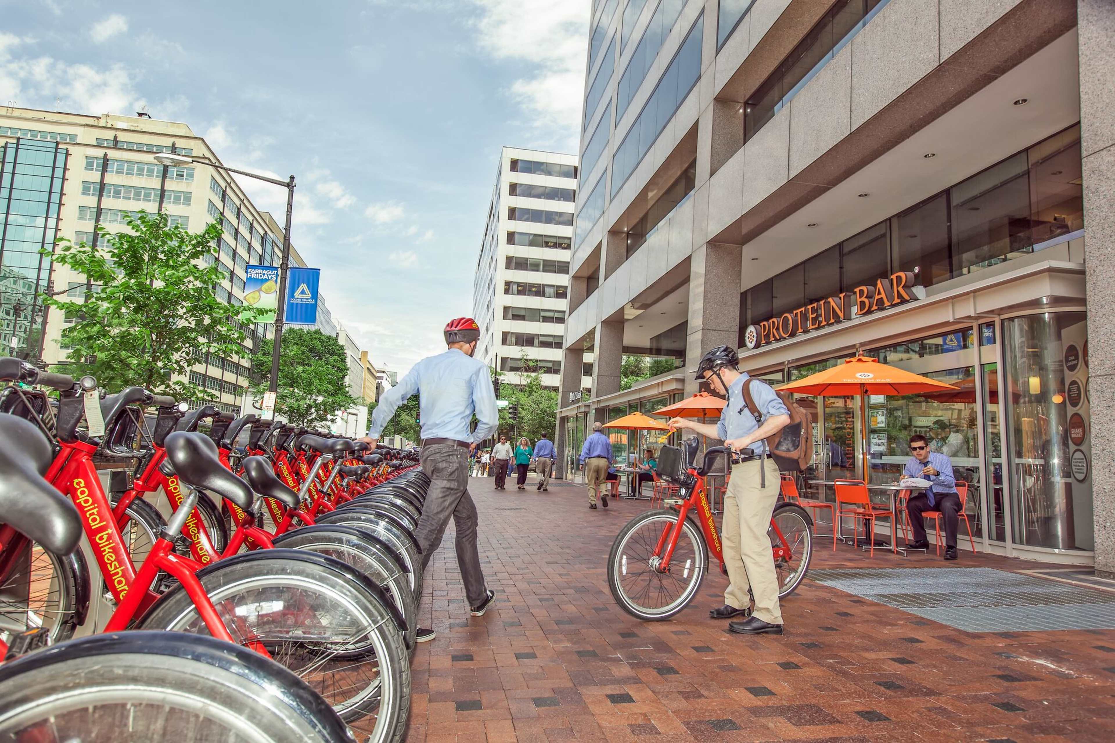 A row of rental bikes is lined up along a sidewalk in front of a modern building with an outdoor dining area for a restaurant.