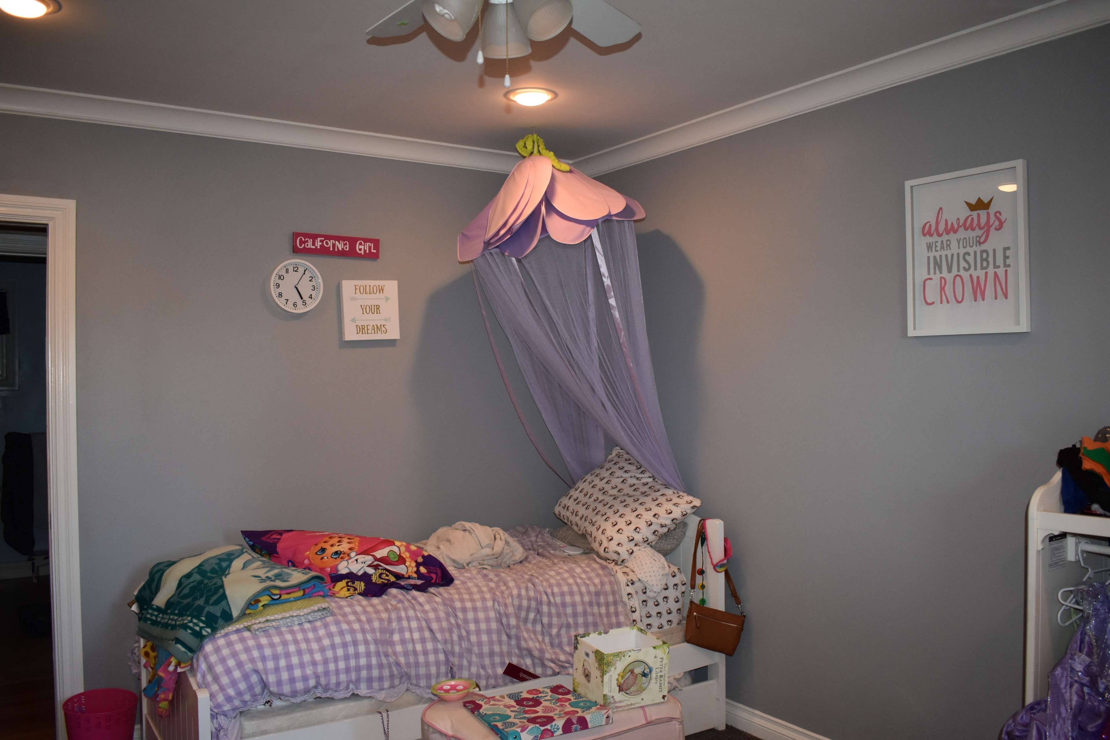 A bedroom features a bed with a purple canopy, a clock on the wall, and various colorful blankets and pillows scattered on the bed.