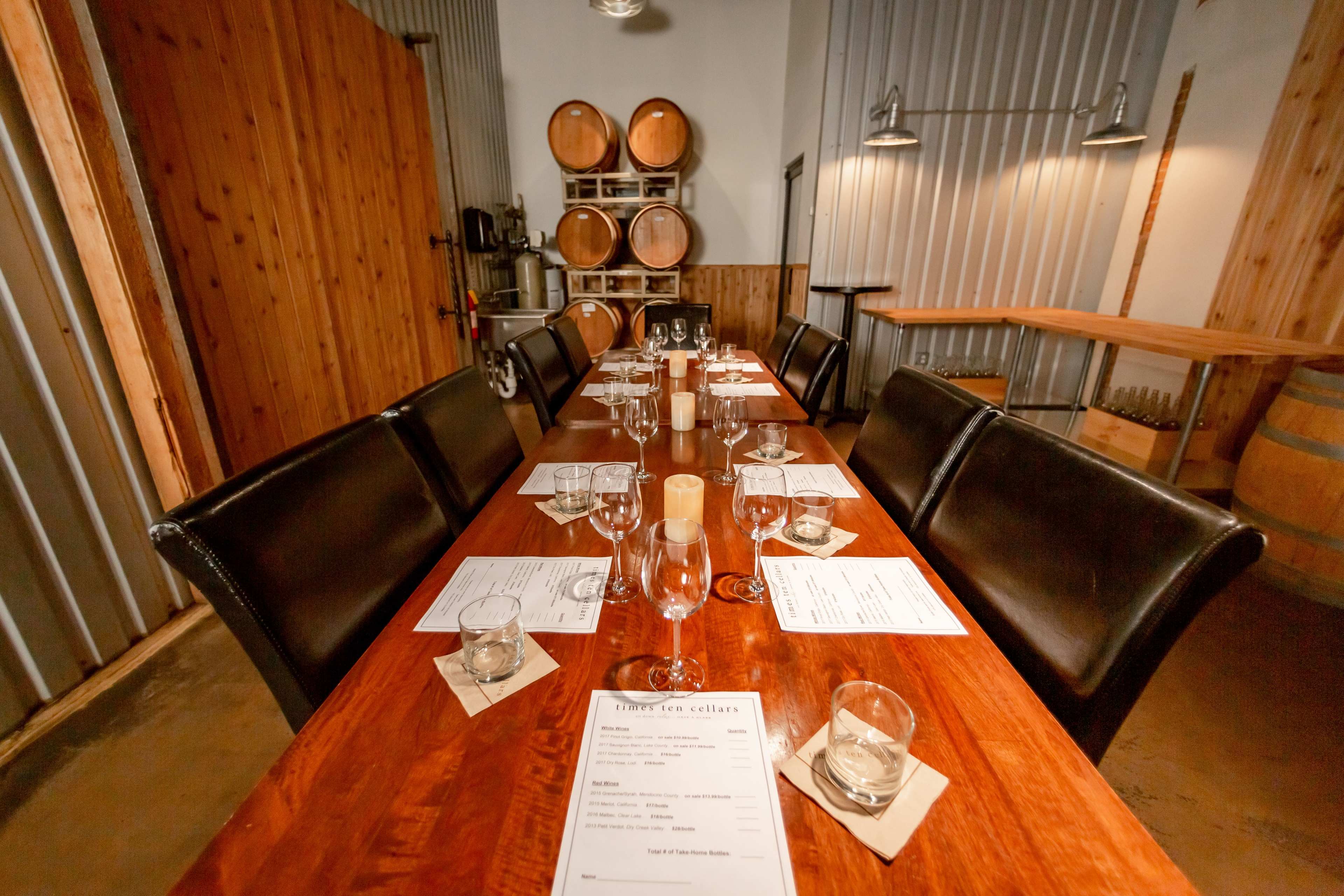 A long wooden dining table is set with glasses and menus in a room featuring wooden barrels and metal walls.