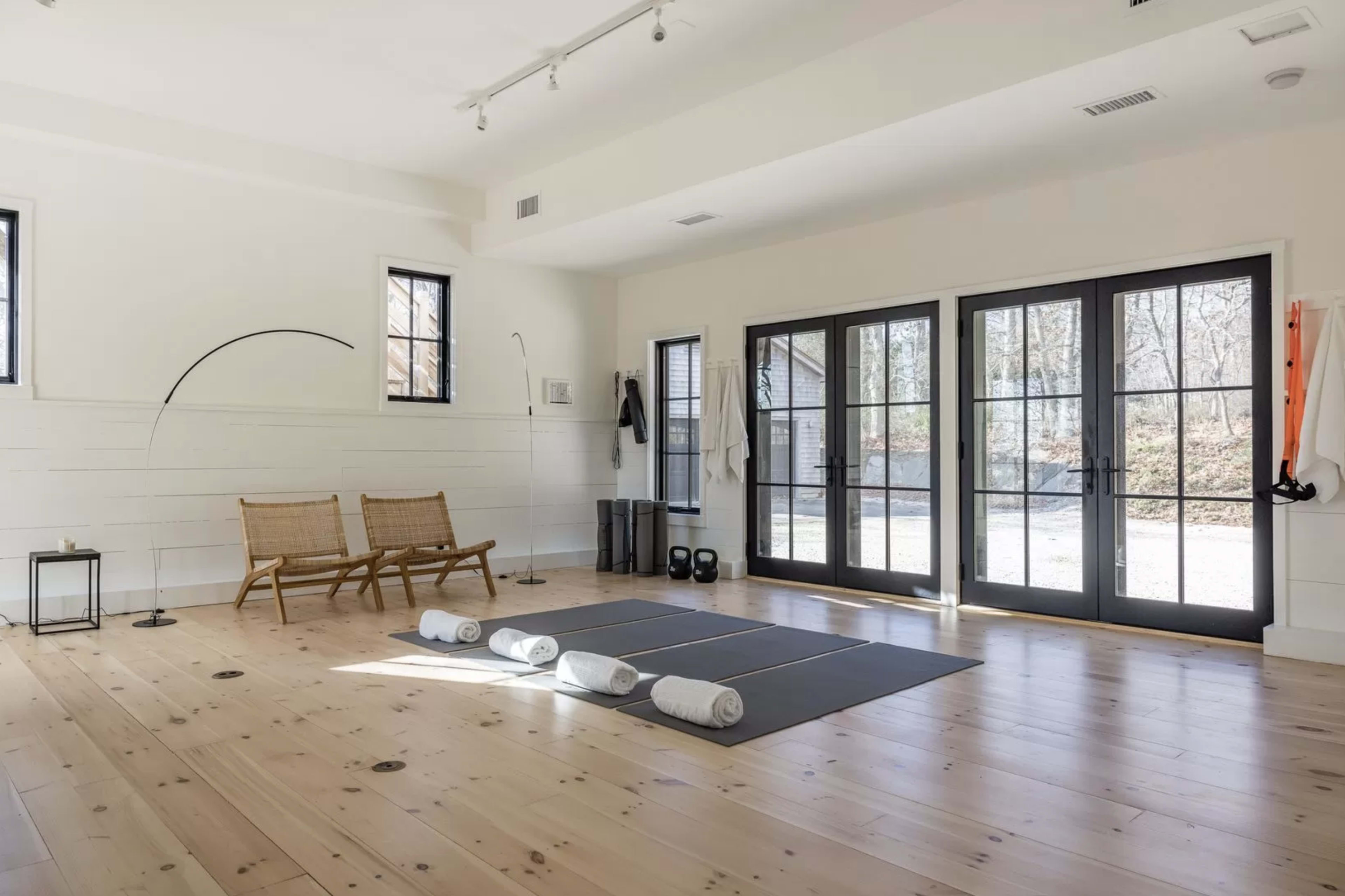A light-filled indoor space designed for yoga, featuring a yoga mat, rolled towels, a pair of chairs, and large glass doors opening to a view outside.