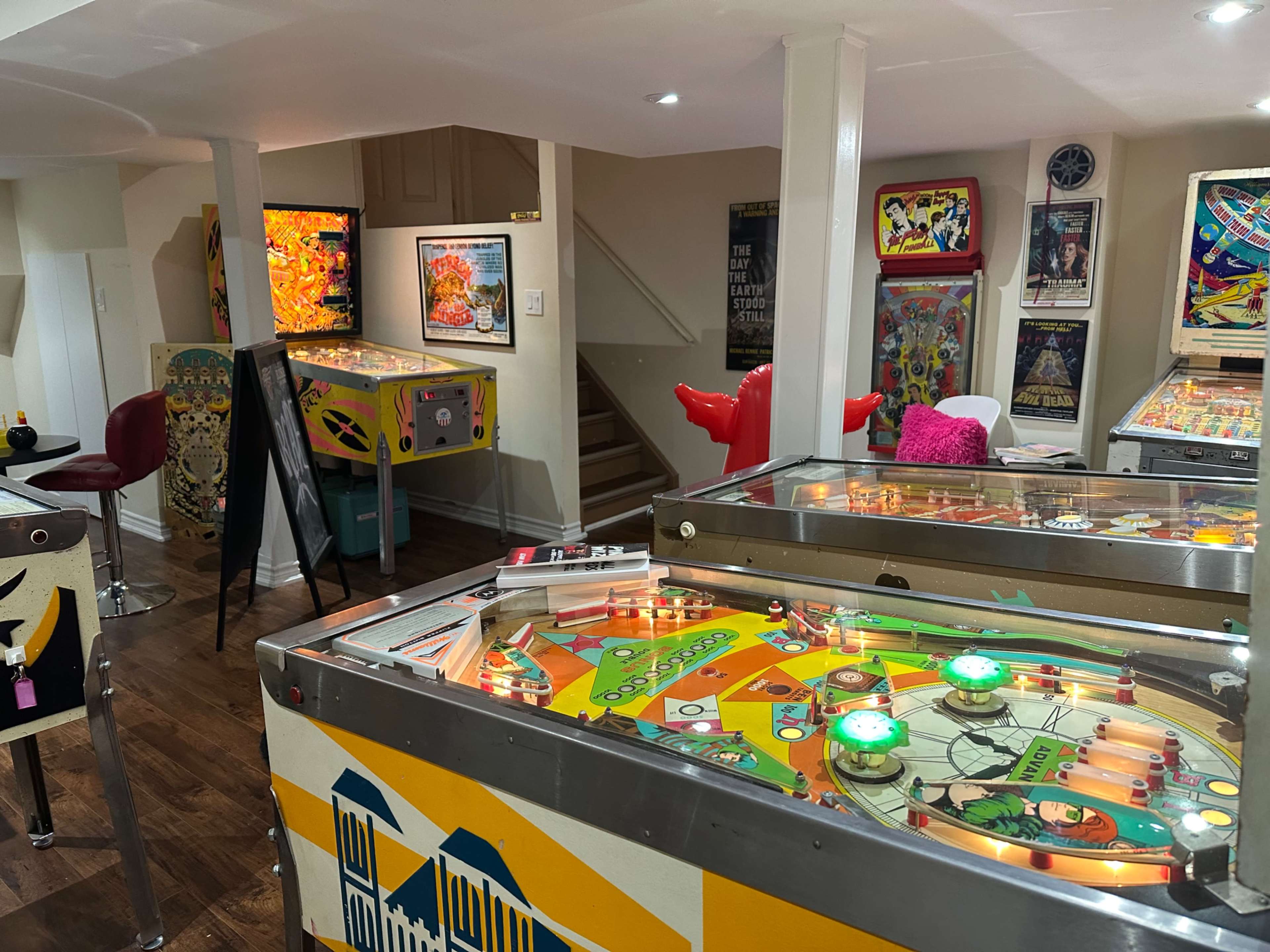 The image shows a gaming room filled with several vintage pinball machines and arcade games arranged in a brightly lit basement.
