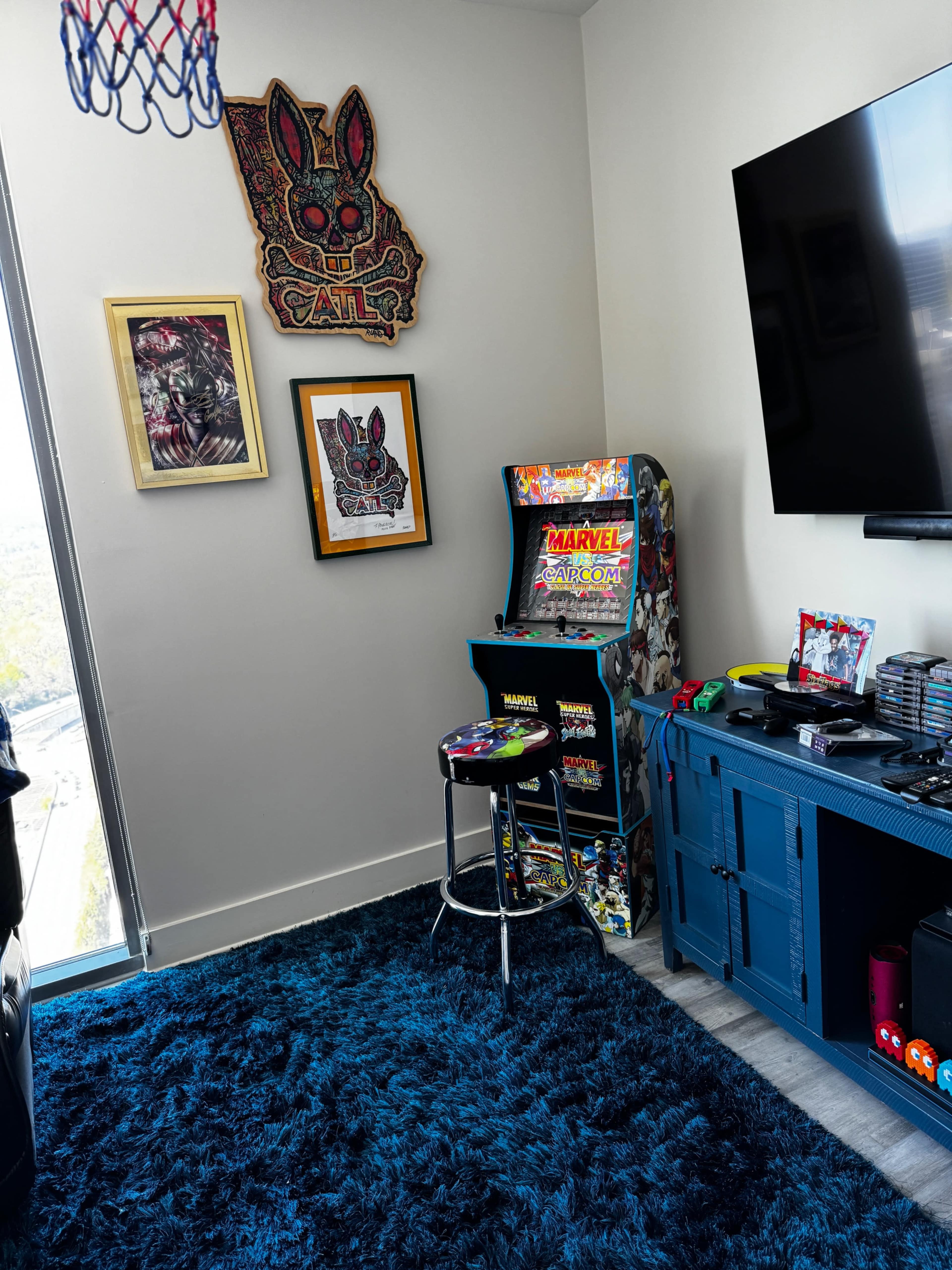 The image shows a corner of a room featuring a Marvel arcade game, wall art, and a plush blue carpet.