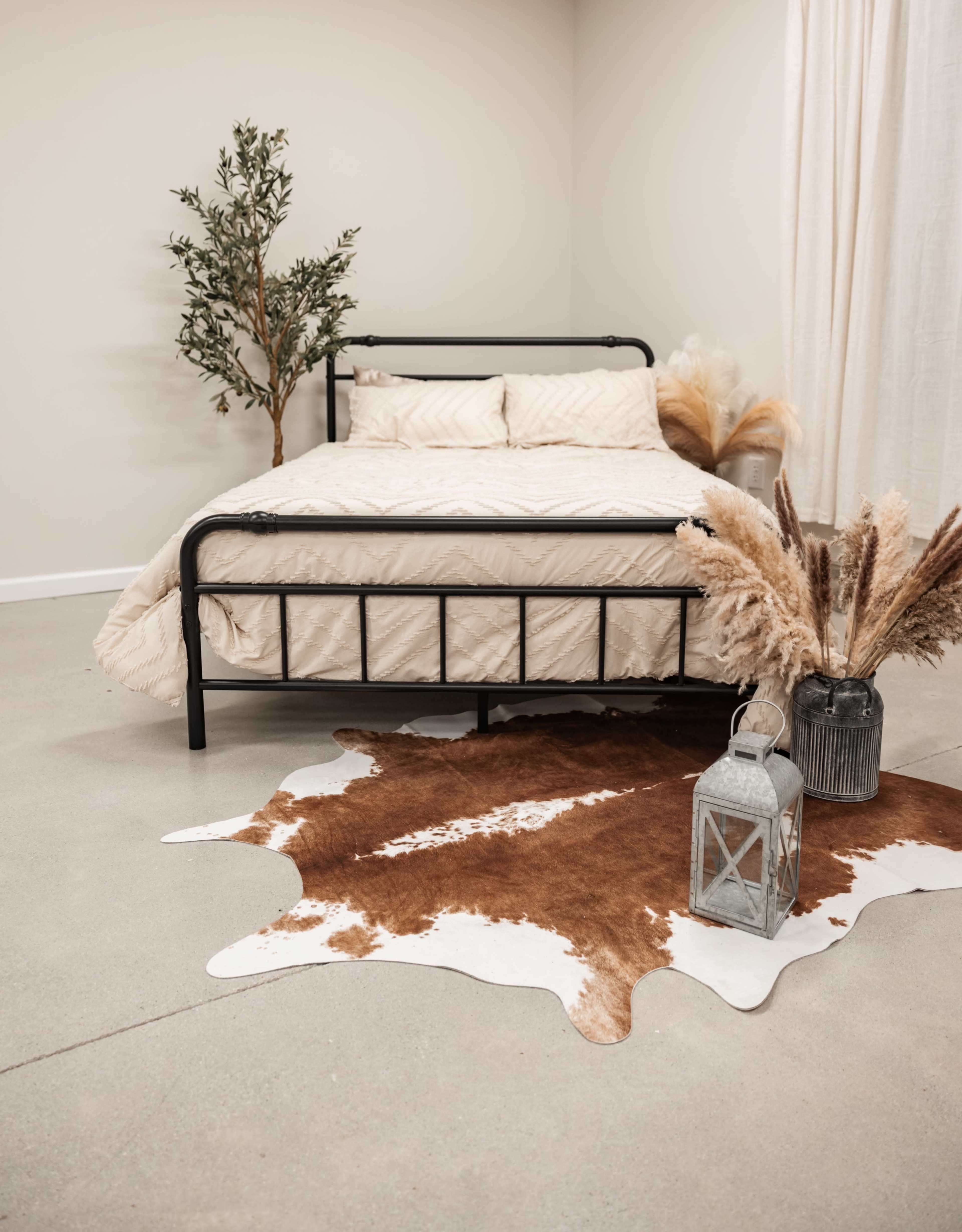 A metal bed with a patterned beige comforter is positioned on a concrete floor, surrounded by a cowhide rug, a lantern, and decorative plants.