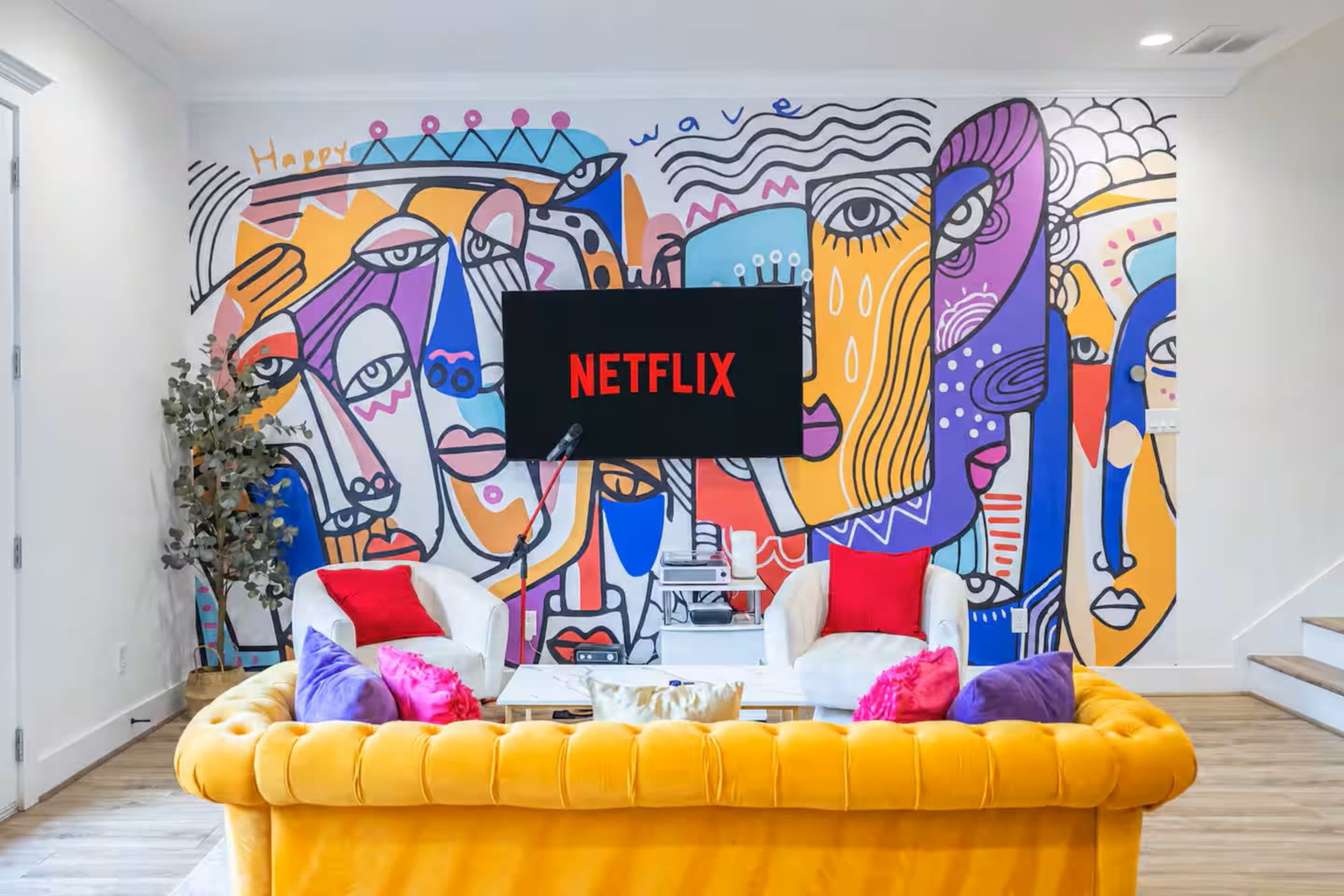 The image shows a vibrant living room with a yellow couch, colorful artwork featuring faces on the wall, and a television displaying the Netflix logo.