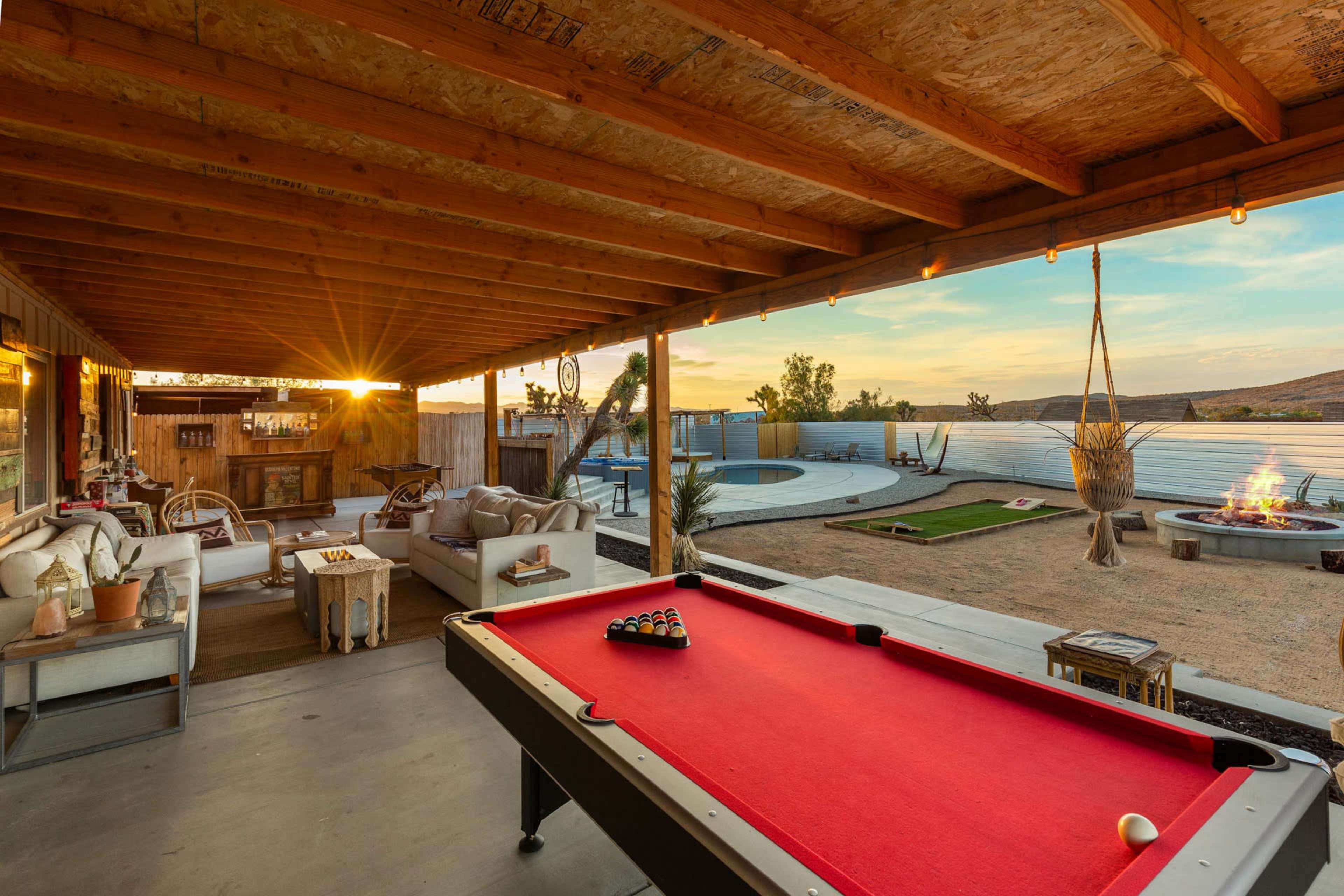 The image shows a covered outdoor space featuring a red pool table, comfortable seating, and a view of a fire pit and swimming pool in a desert landscape at sunset.
