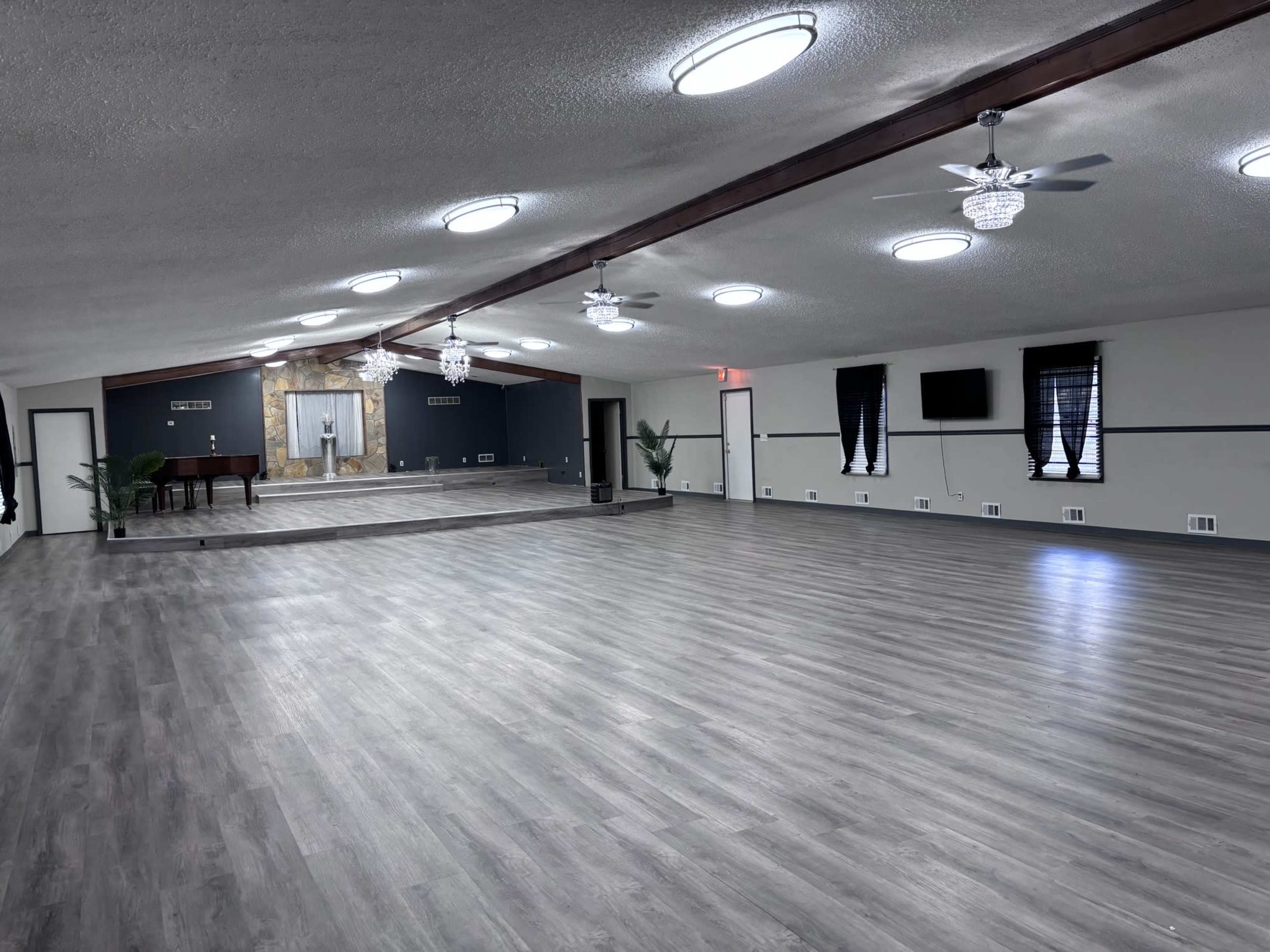 The image shows a spacious, empty interior with grey wooden flooring, modern lighting, and a mirrored stage at one end.