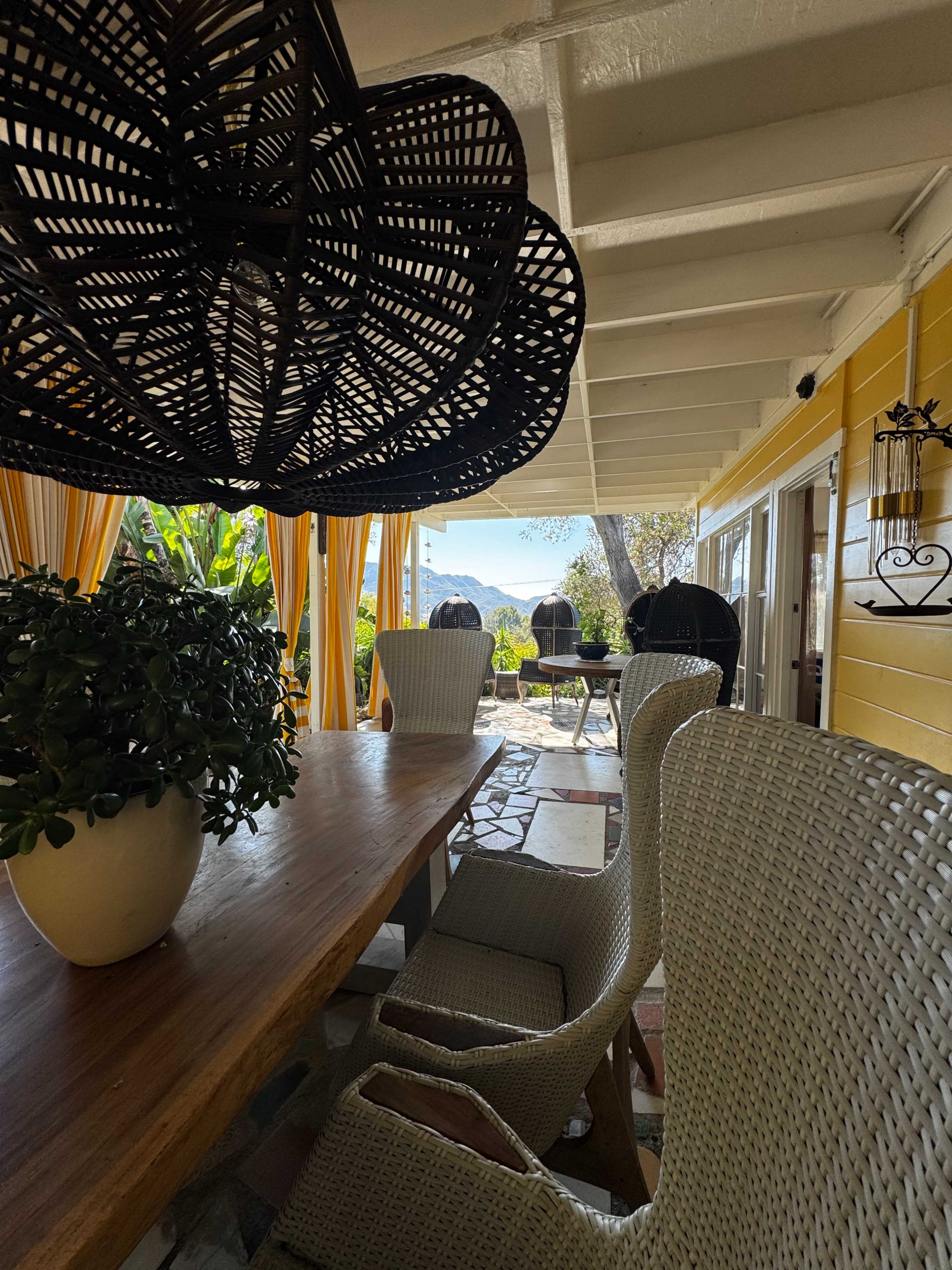 The image shows a brightly colored outdoor dining area with a wooden table and wicker chairs, framed by a view of lush greenery and mountains beyond.
