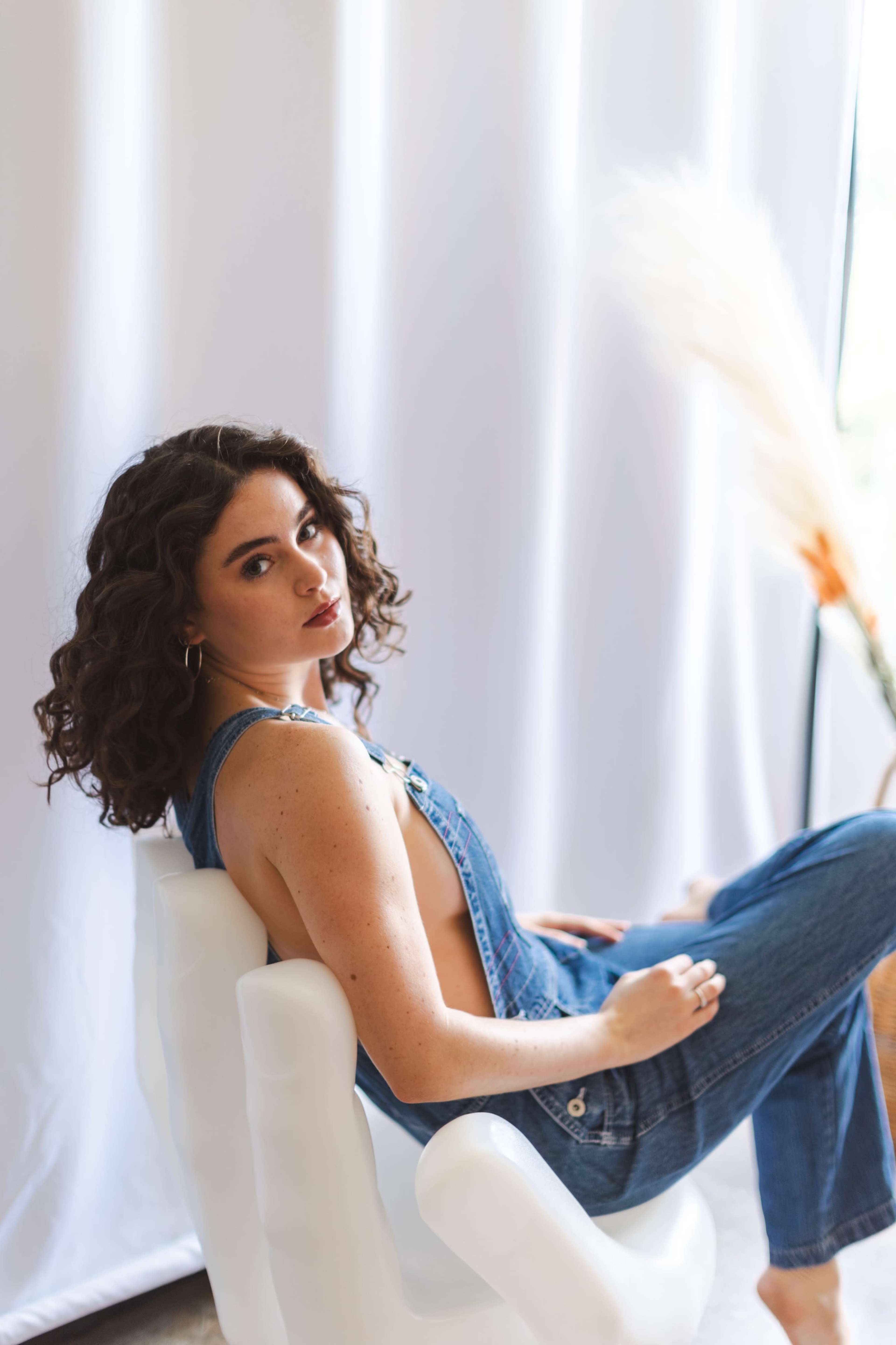 A woman with curly hair wearing overalls sits on a white chair in front of a white backdrop.