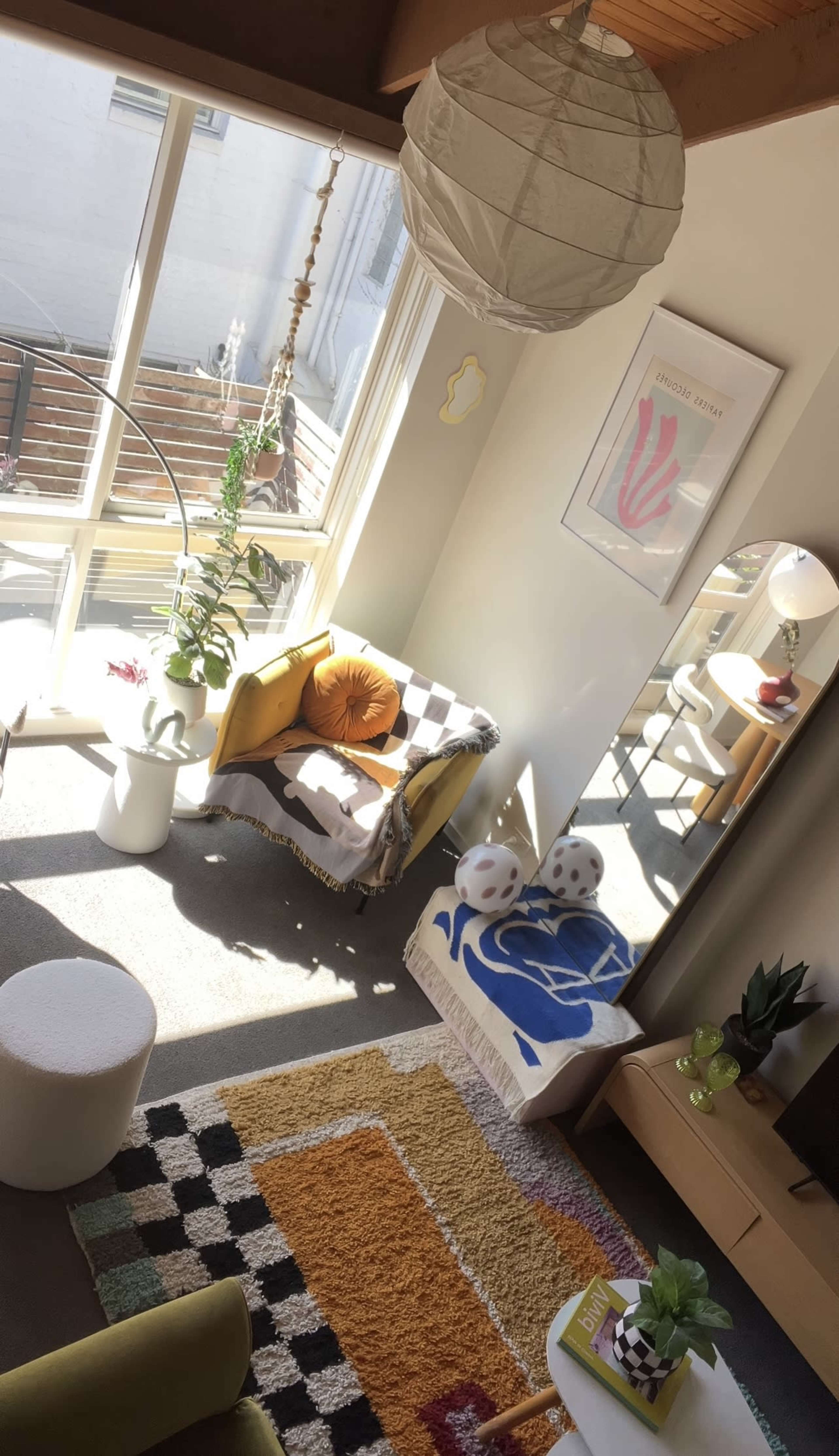 A sunlit living room features a yellow armchair and a large mirror, with decorative plants, a colorful rug, and a small coffee table.