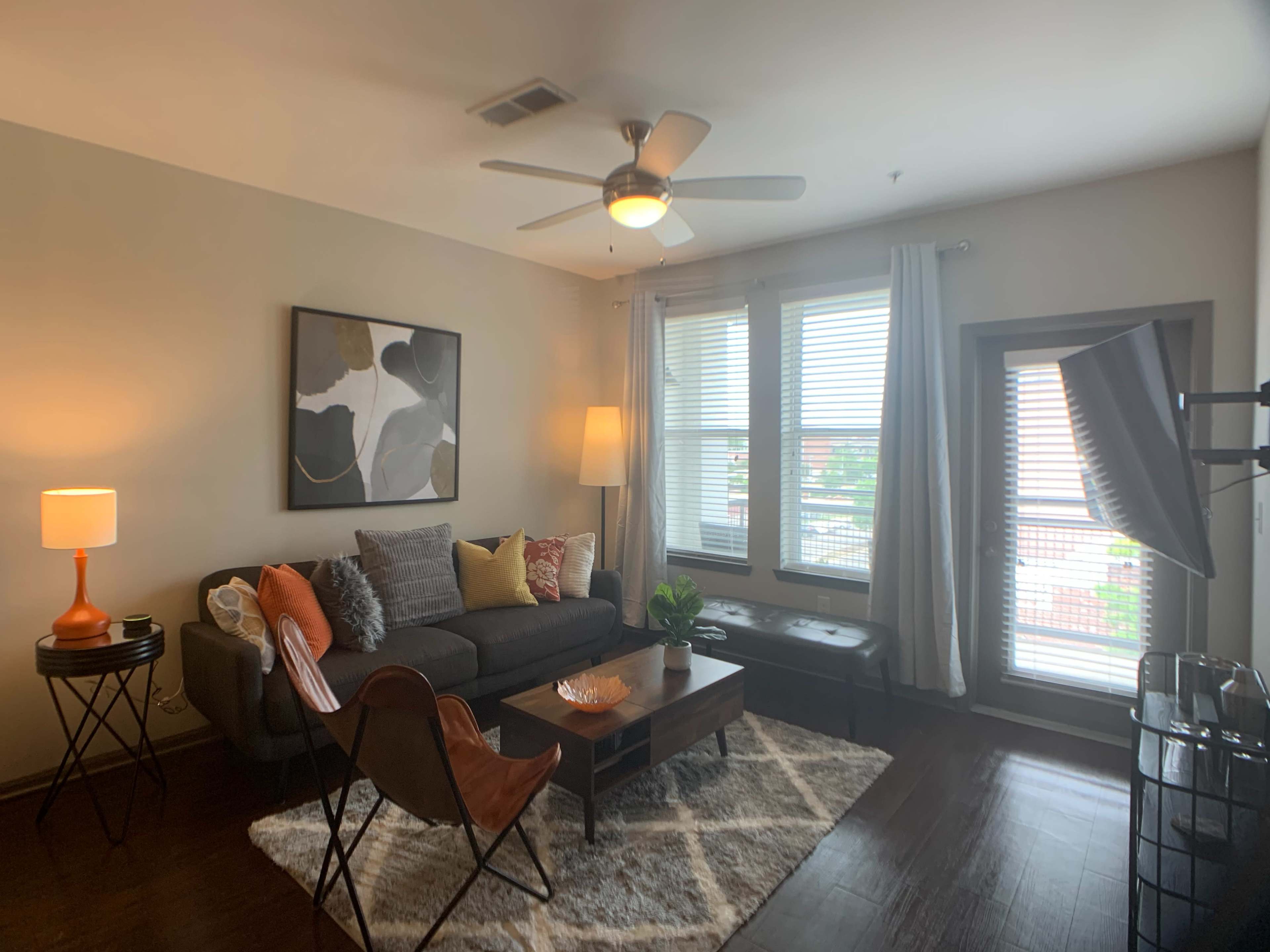 A neatly arranged living room featuring a dark gray sofa with decorative pillows, a coffee table, a modern chair, a lamp, and large windows with sheer curtains.