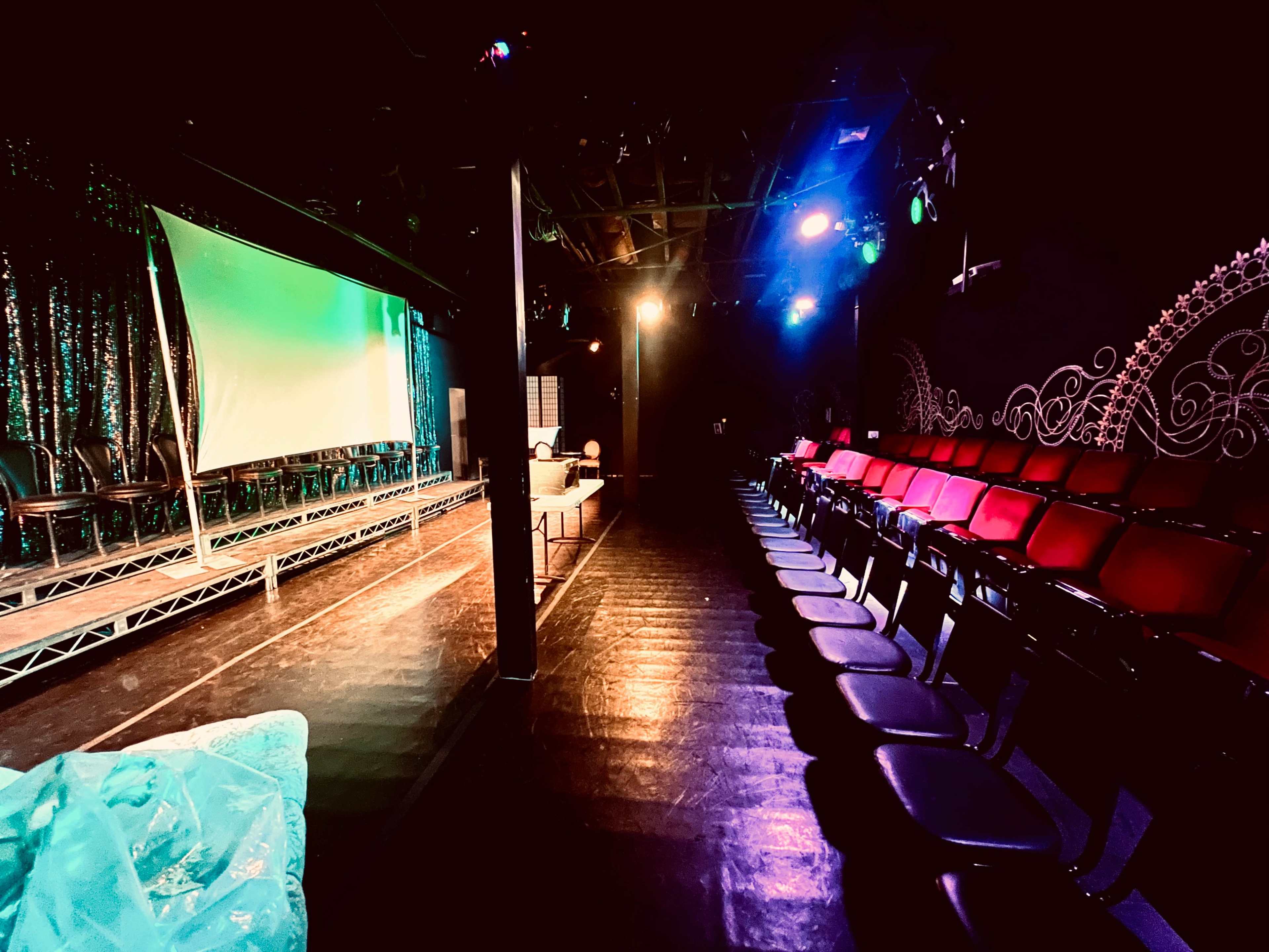 A small theater setting with a stage in the foreground, illuminated by colorful lights, and rows of empty chairs facing the stage.