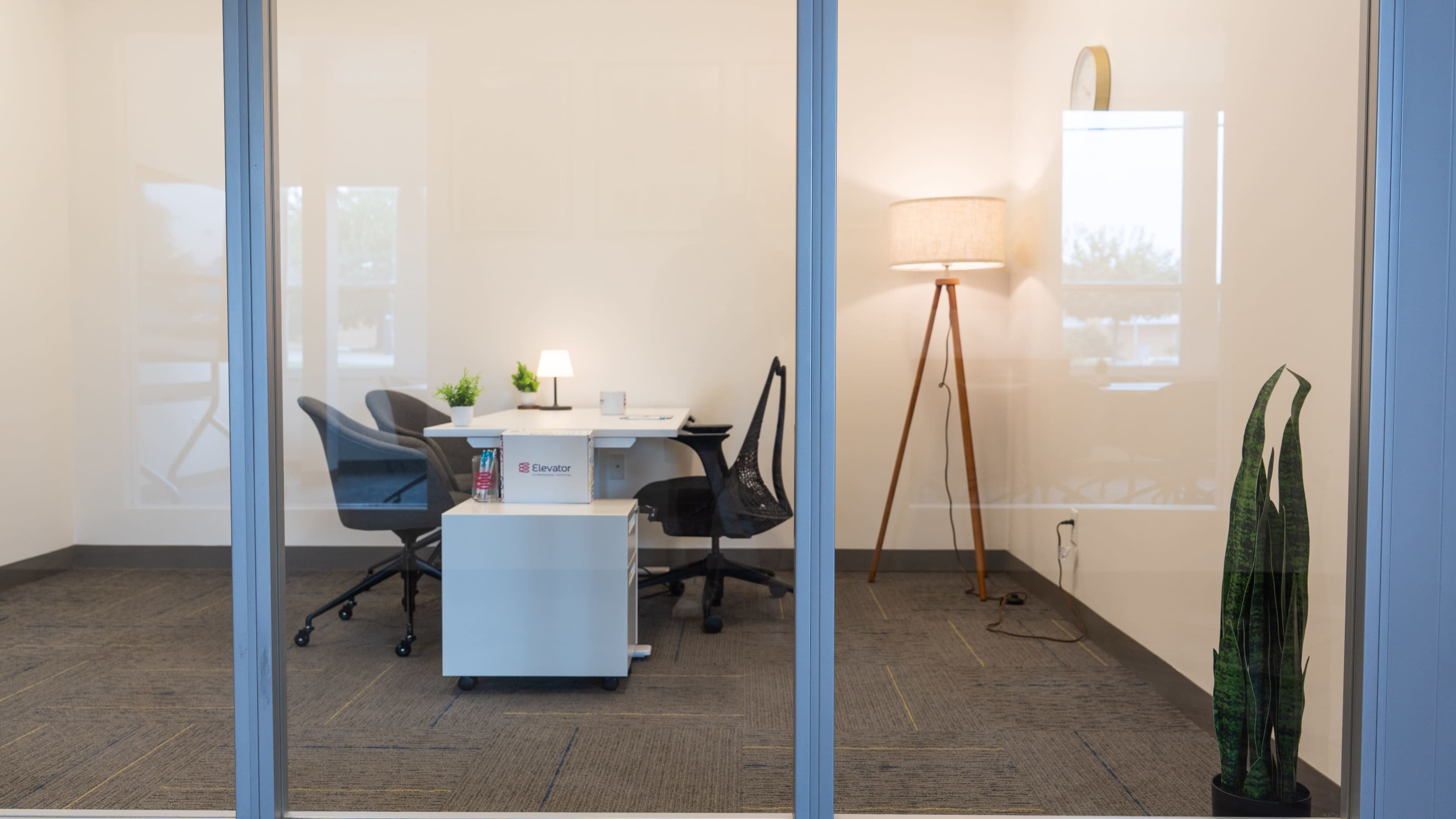 The image shows a modern office space with a table, four chairs, a lamp, and a potted plant, all visible through glass walls.