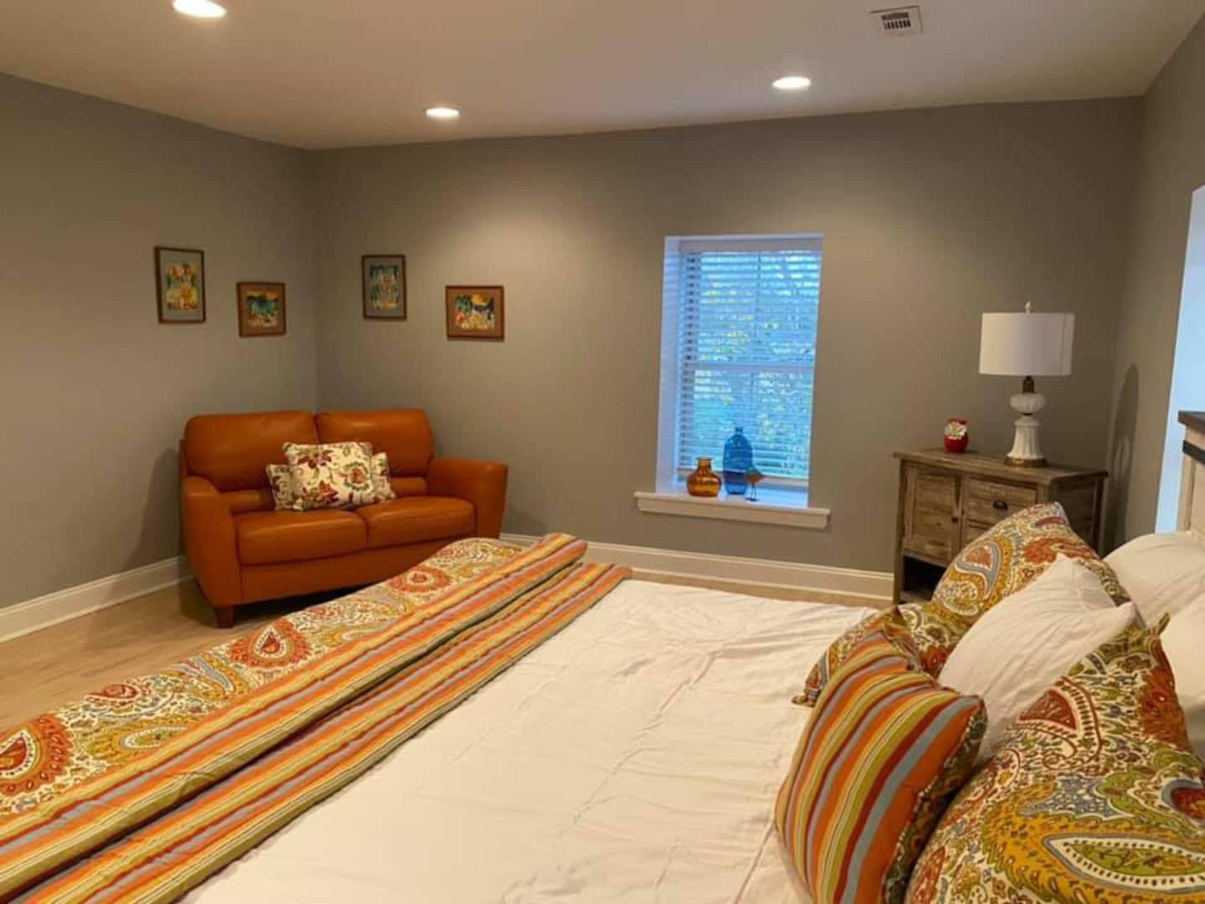 A bedroom featuring a brown sofa with decorative pillows, a bed with colorful patterned bedding, and a window with light filtering through.