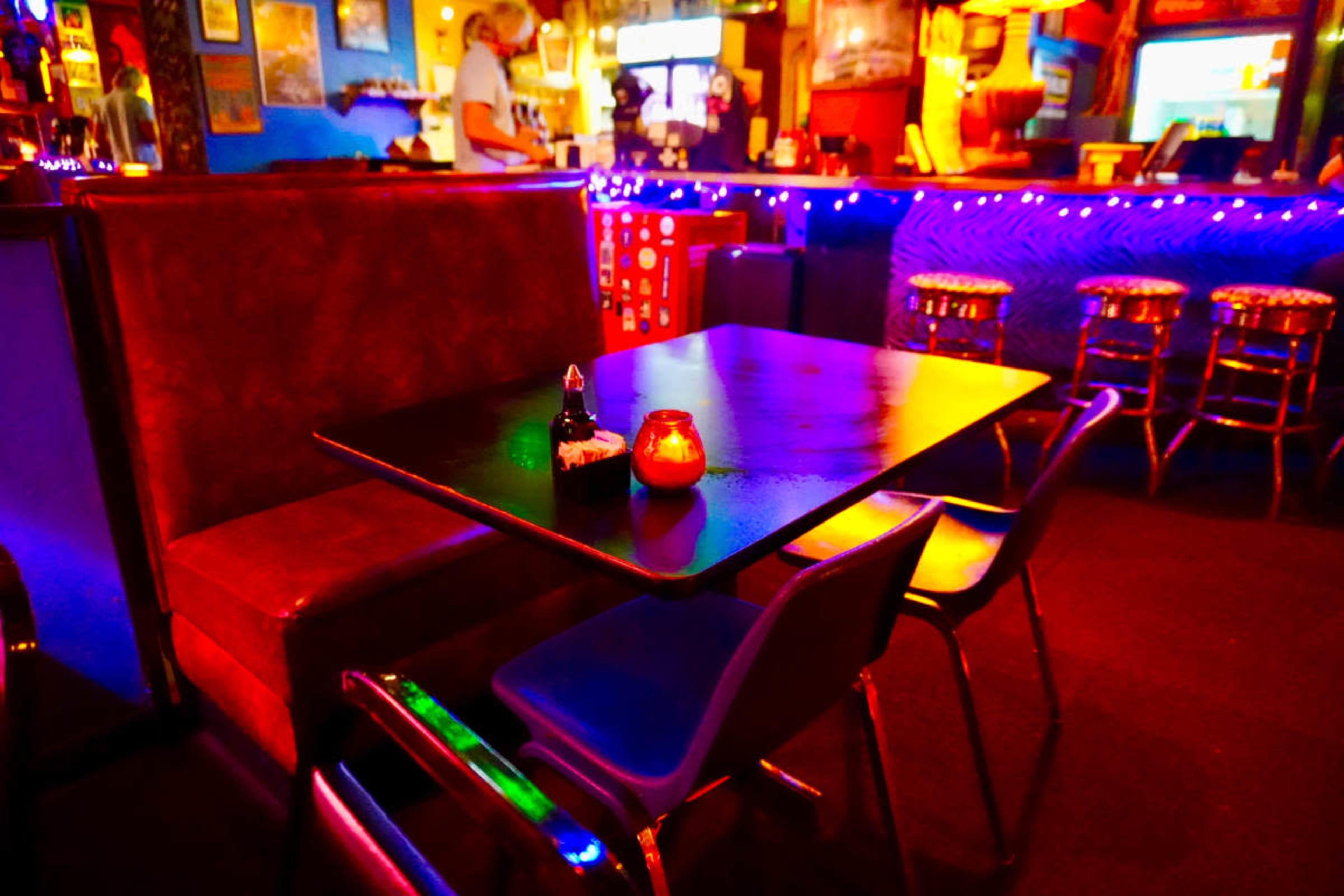 The image shows a dimly lit bar with colorful neon lights, a booth seating area, and a table set with condiments and candles.