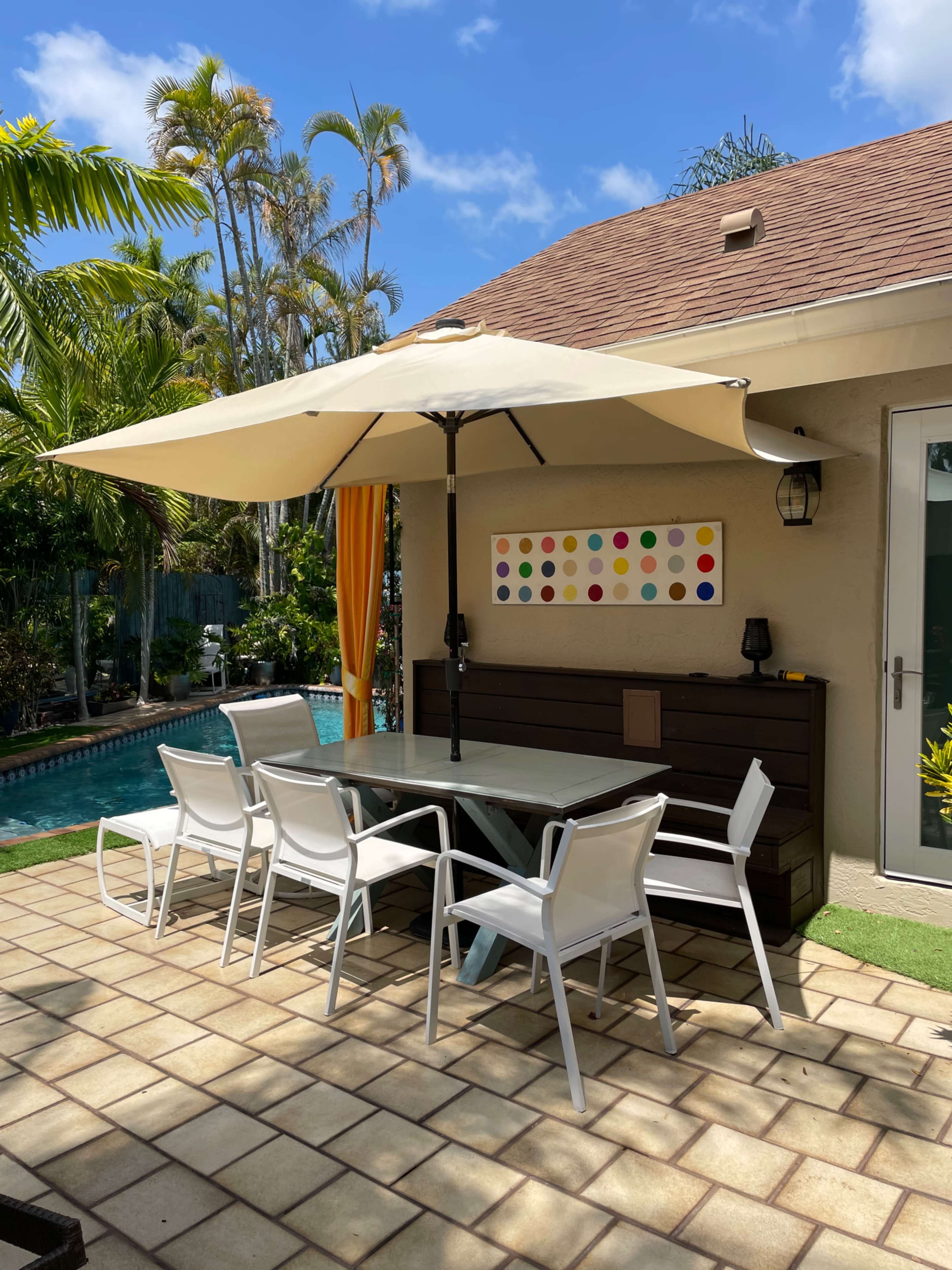 A patio area features a large umbrella over a rectangular table surrounded by white chairs, with a pool and palm trees in the background.
