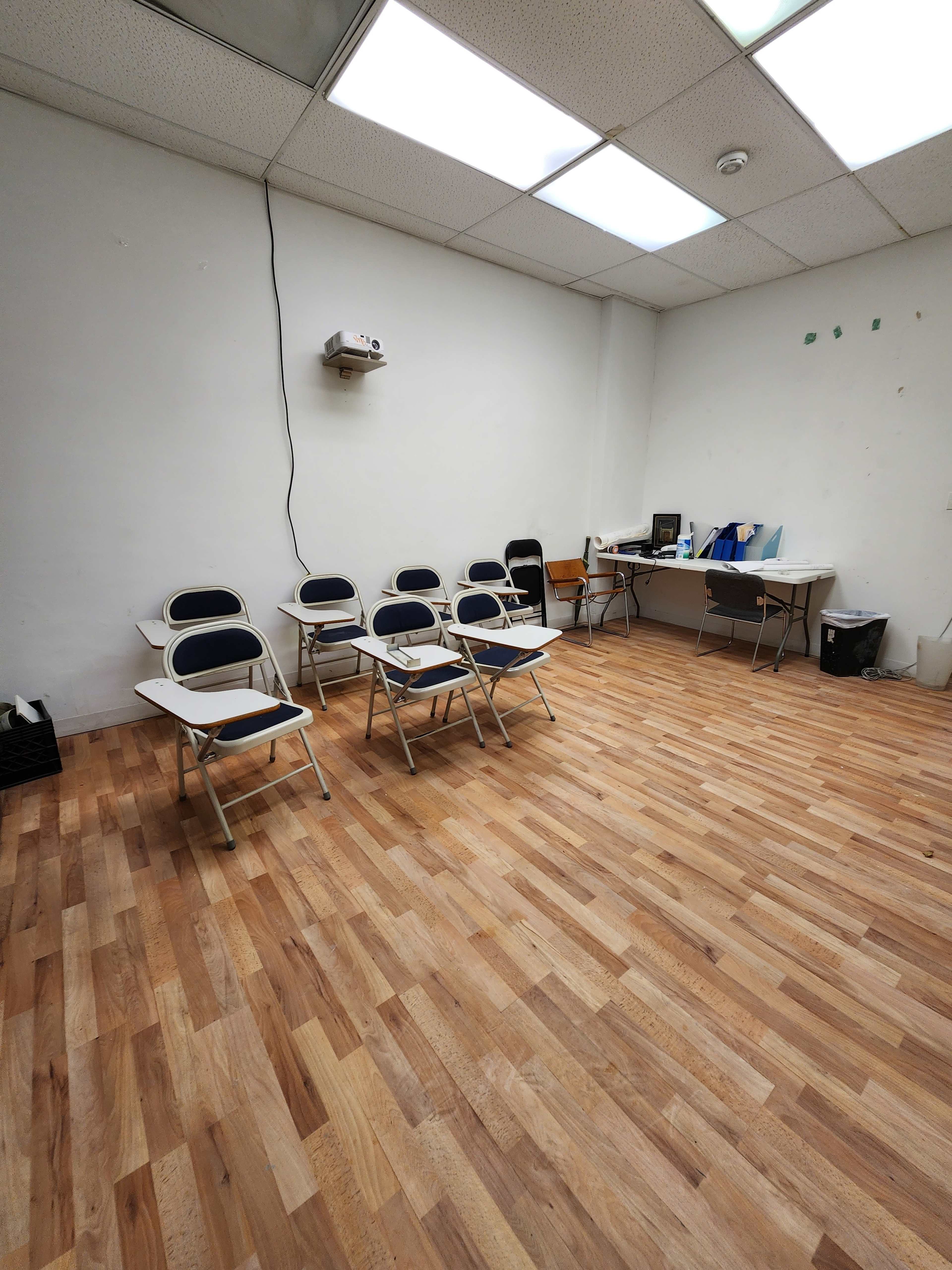 A sparsely furnished room with a wood laminate floor, several metal chairs arranged in a row, and a desk equipped with a computer and a projector on the wall.
