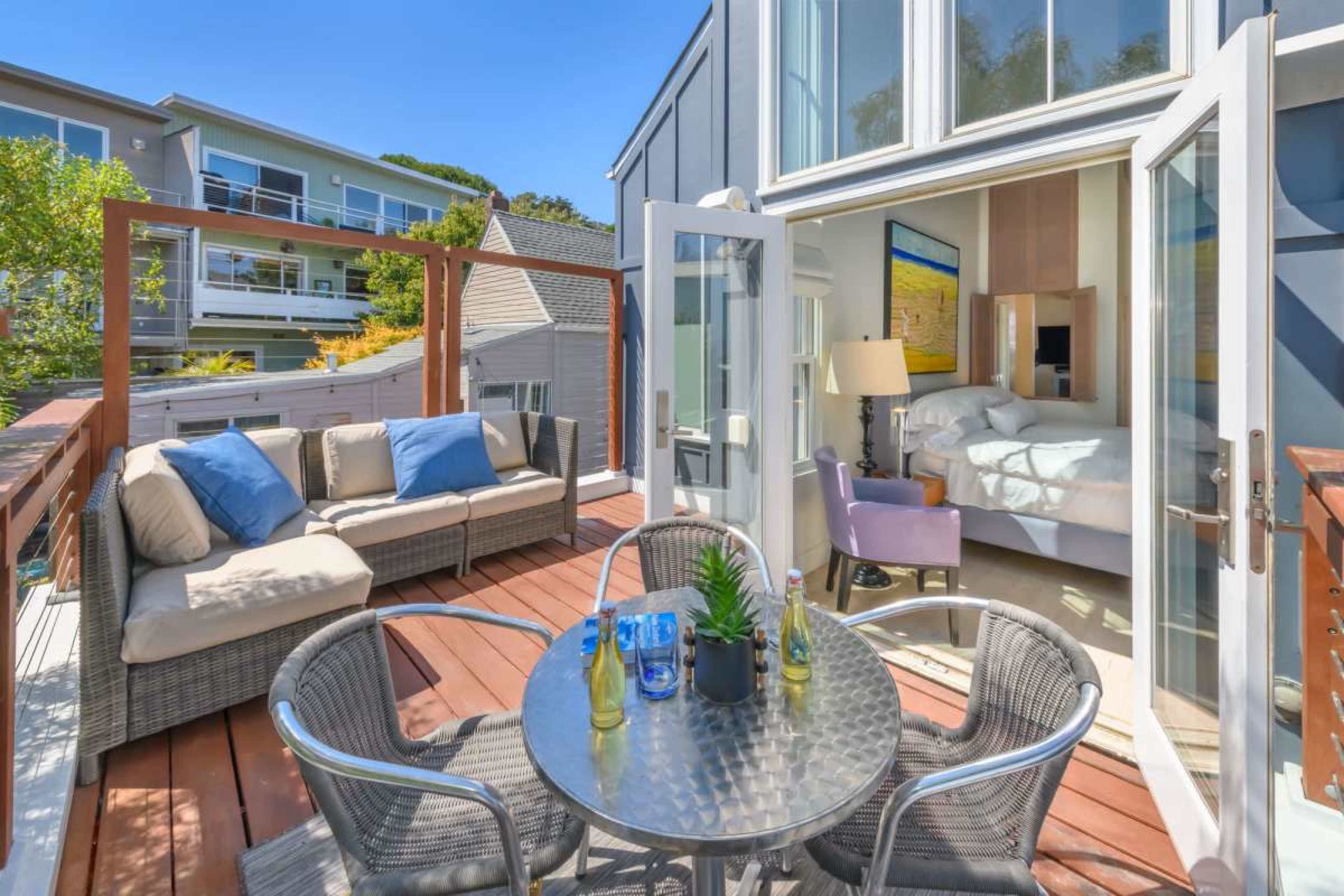 A balcony features outdoor seating, a circular table, and doors opening into a well-lit interior space with a bed.