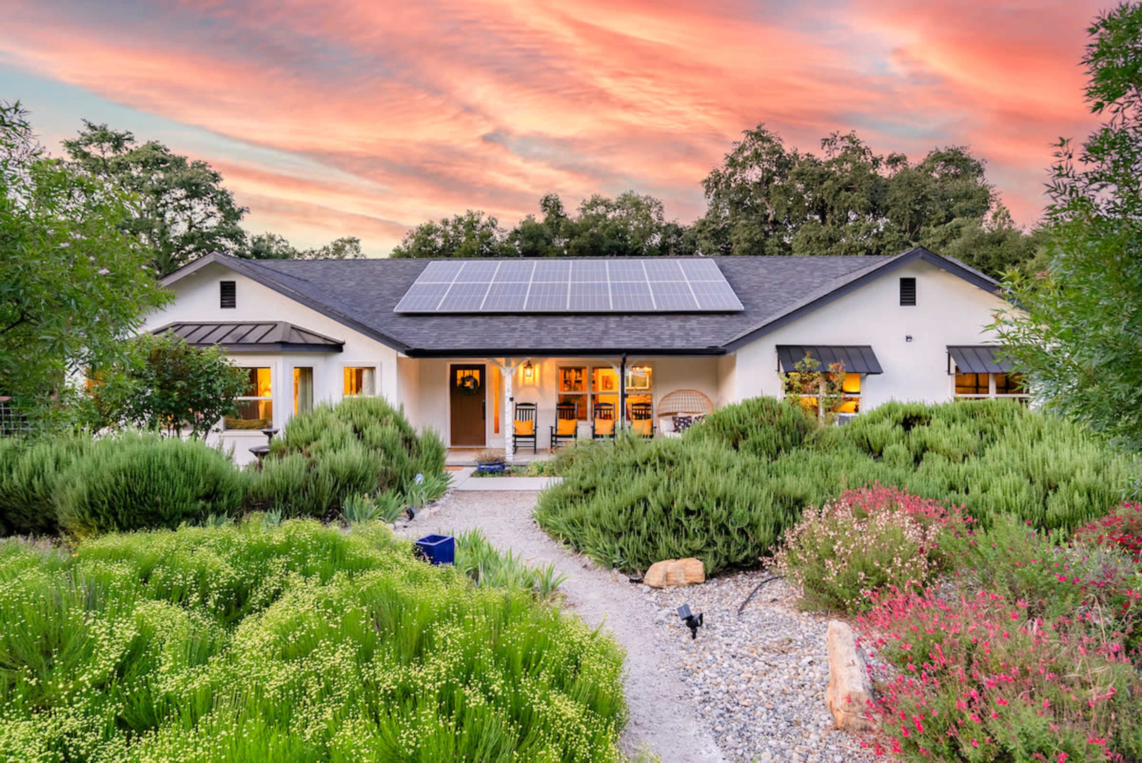 A single-story house with solar panels on the roof is surrounded by a lush, landscaped garden at sunset.
