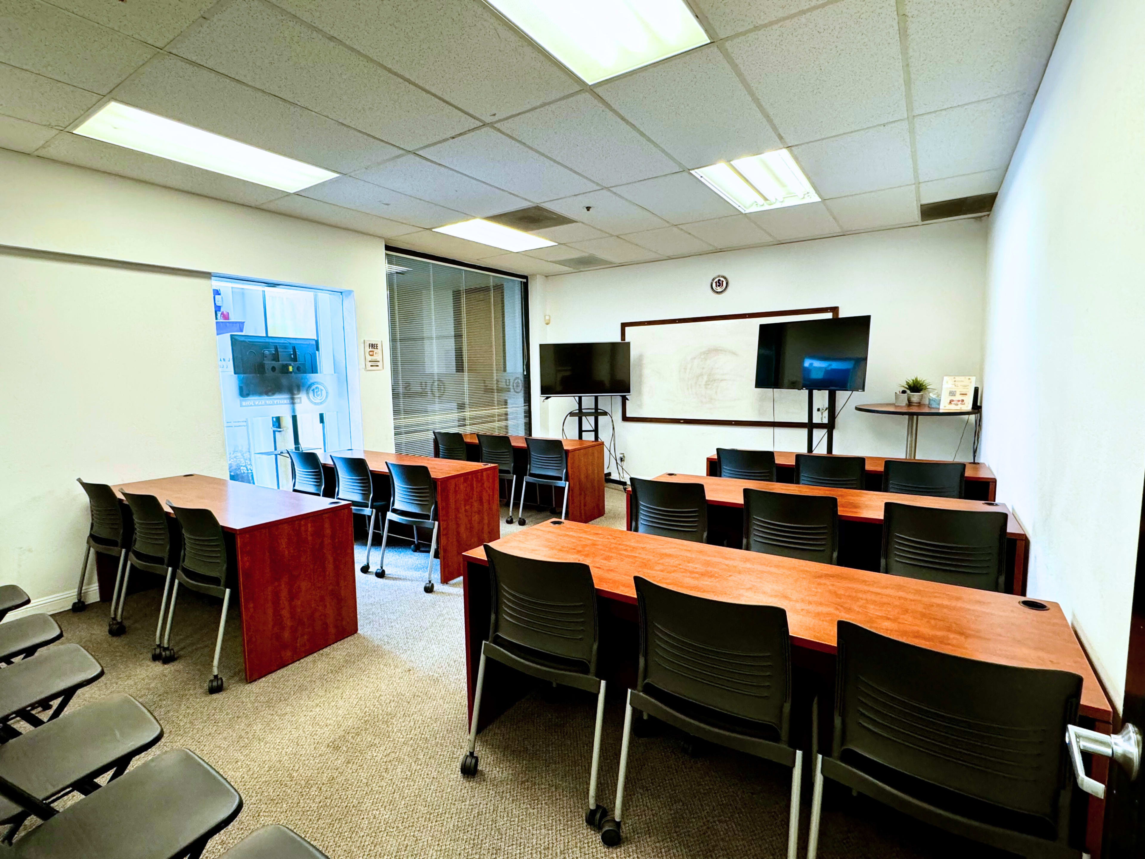 A classroom with rows of tables and chairs, two television screens, and a whiteboard at the front.