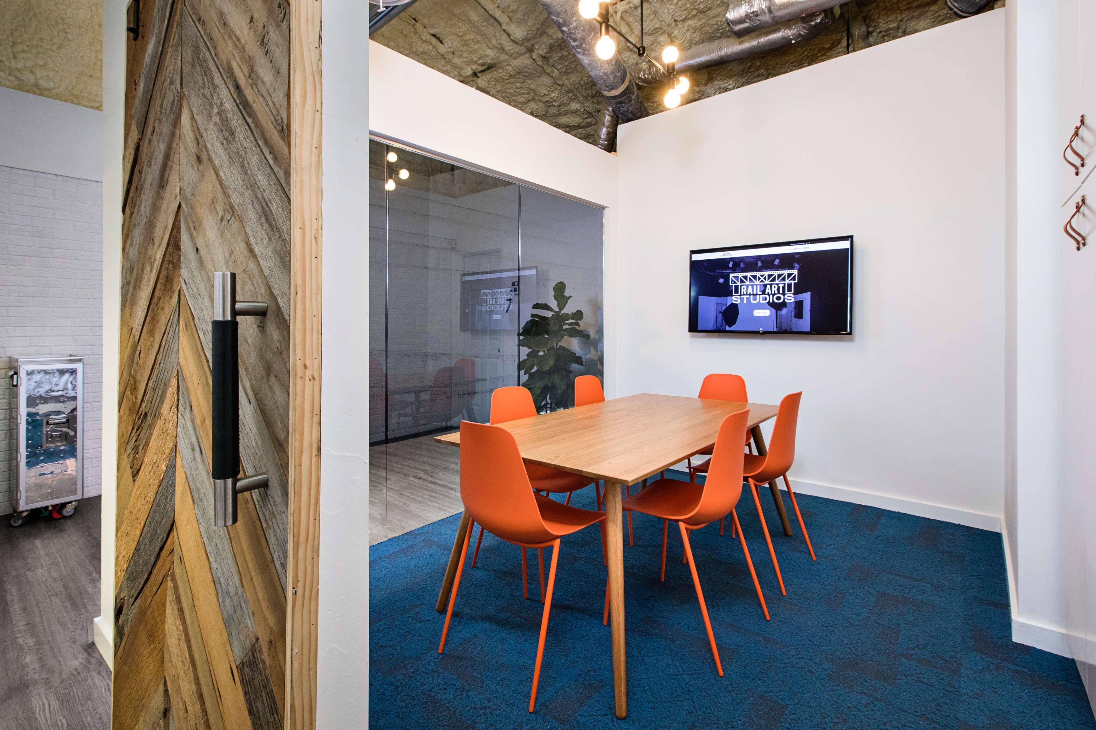 A glass-walled conference room features a wooden table surrounded by orange chairs, with a monitor displaying a graphic on one wall.