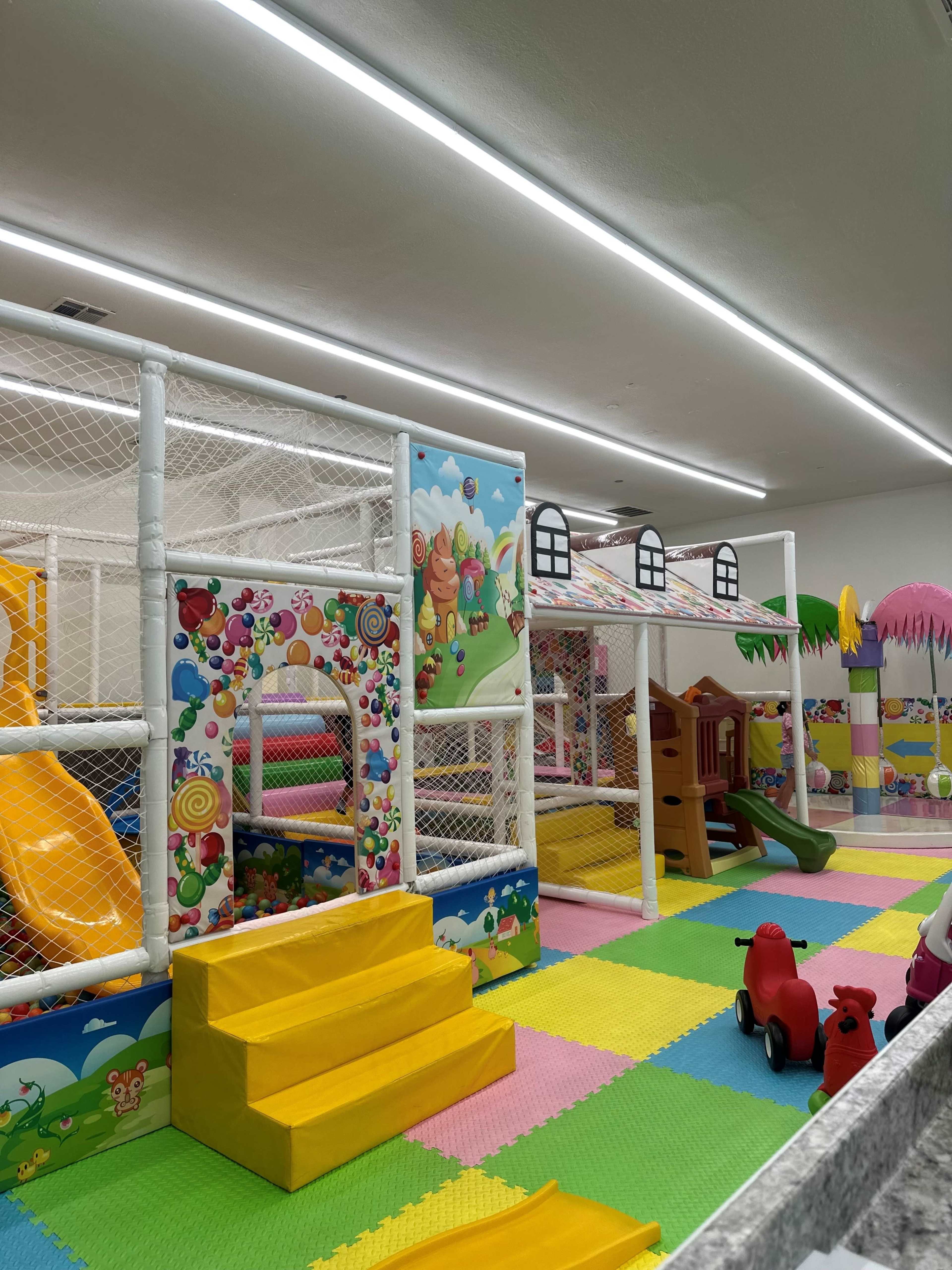 The image shows an indoor play area with colorful mats, climbing structures, slides, and various playful decorations.