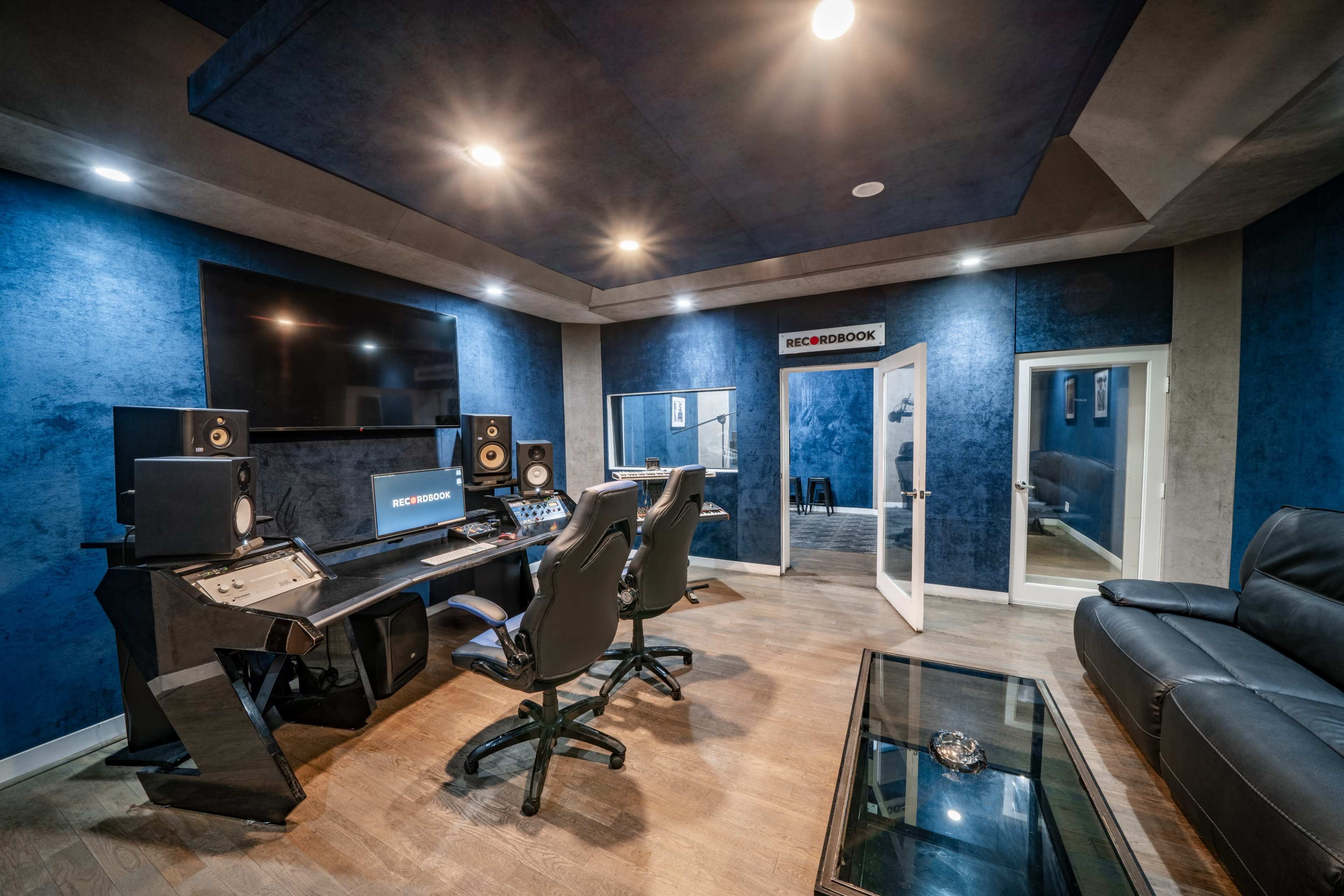 The image shows a modern recording studio with blue walls, a control desk, speakers, and a large monitor, alongside a glass coffee table and a leather sofa.