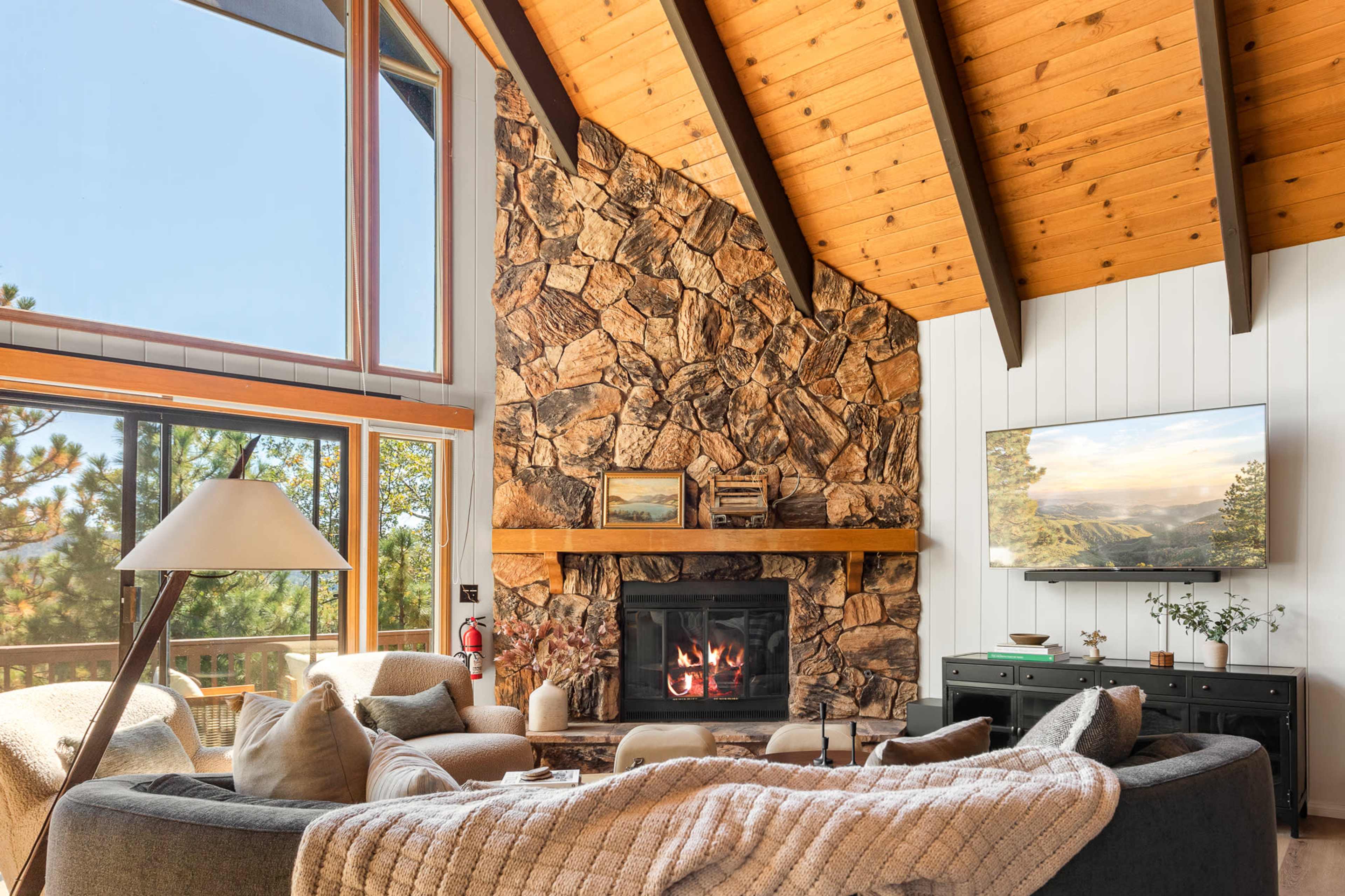 The image shows a cozy living room featuring a large stone fireplace, a sectional sofa, wooden beams on the ceiling, and a flat-screen TV mounted on the wall.