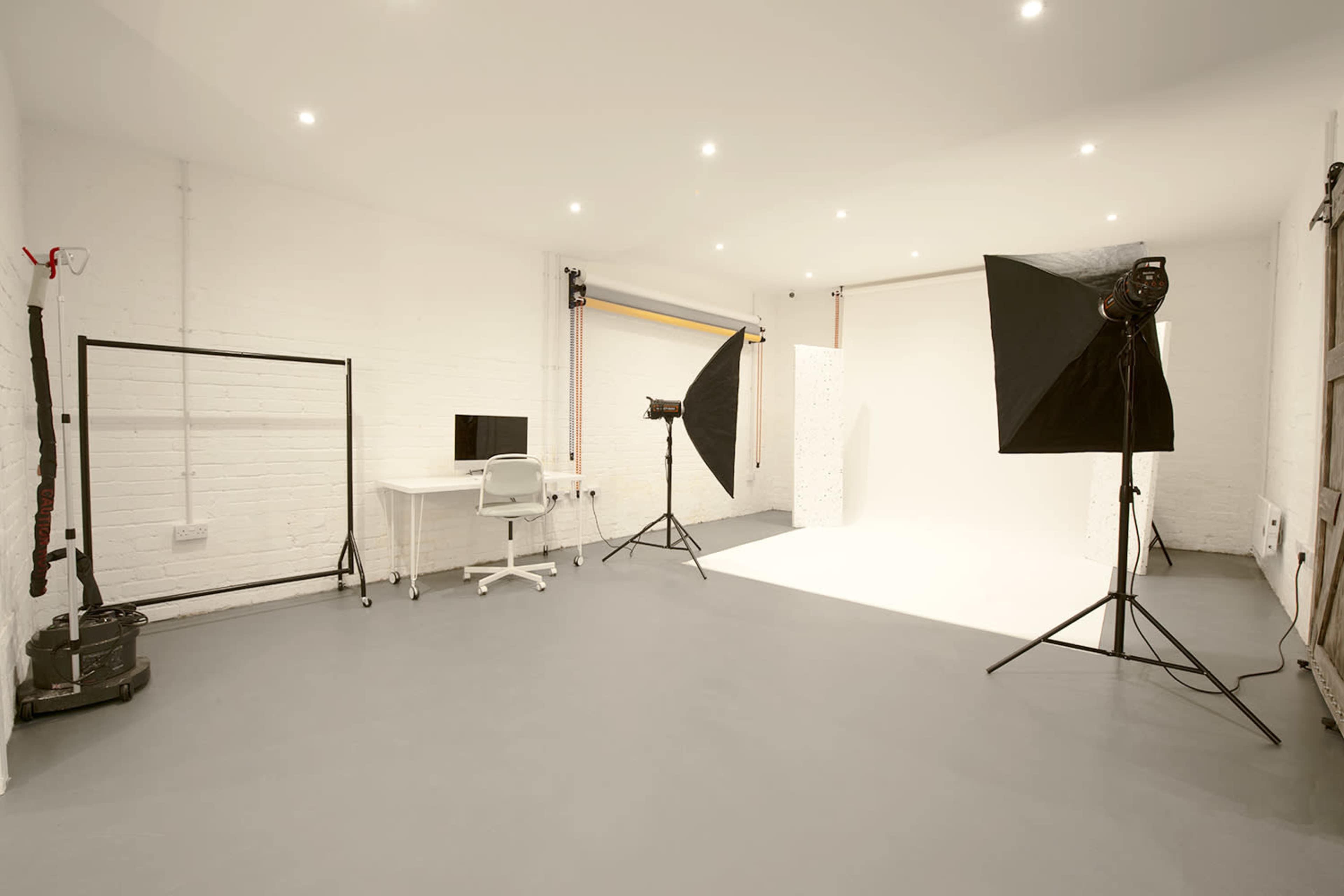 A spacious photography studio with a white backdrop, lighting equipment on stands, a desk with a monitor, and a clean gray floor.