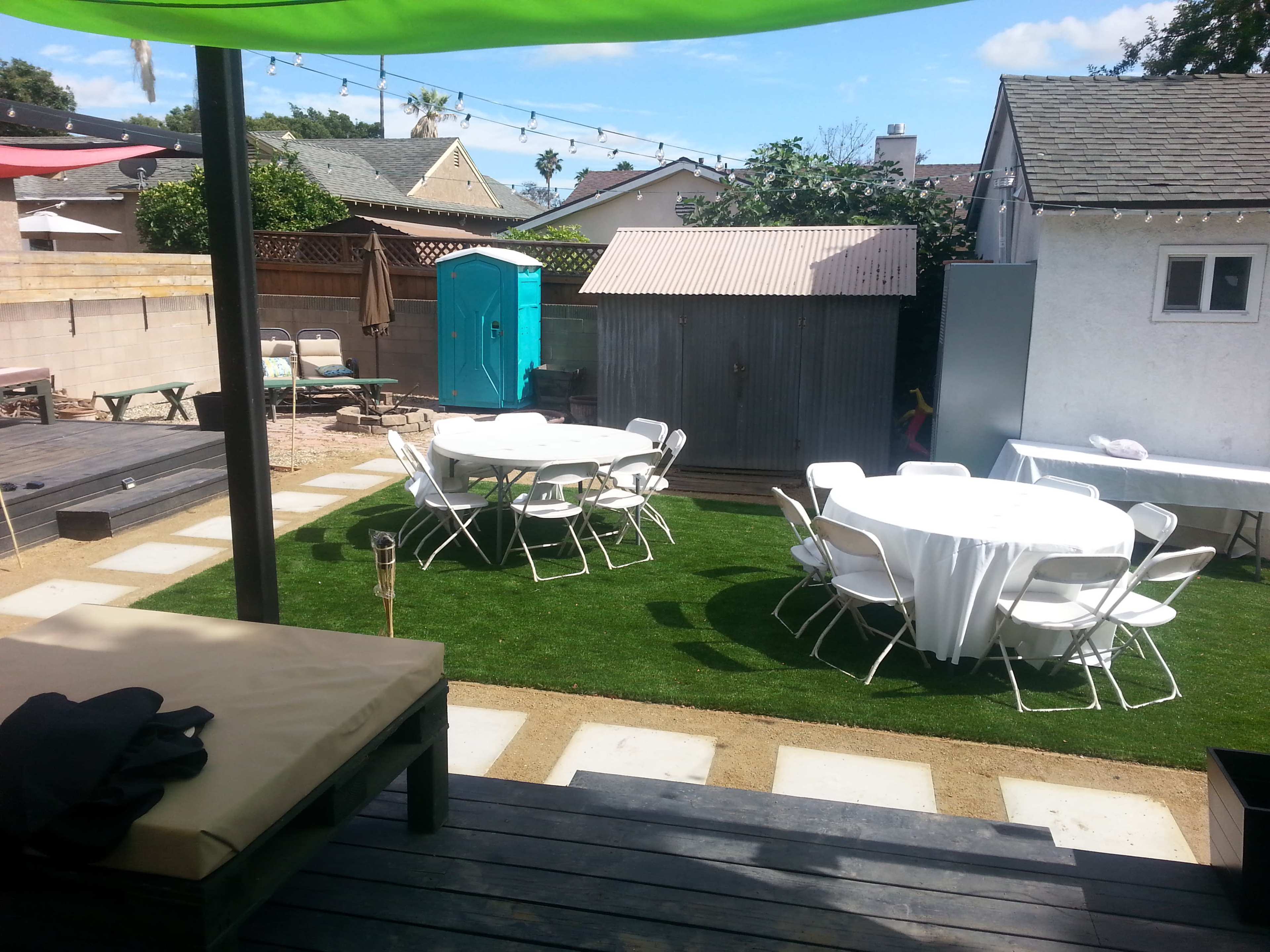 An outdoor space with two round tables covered in white tablecloths, surrounded by white chairs on a grassy area, a shed in the background, and a portable restroom nearby.