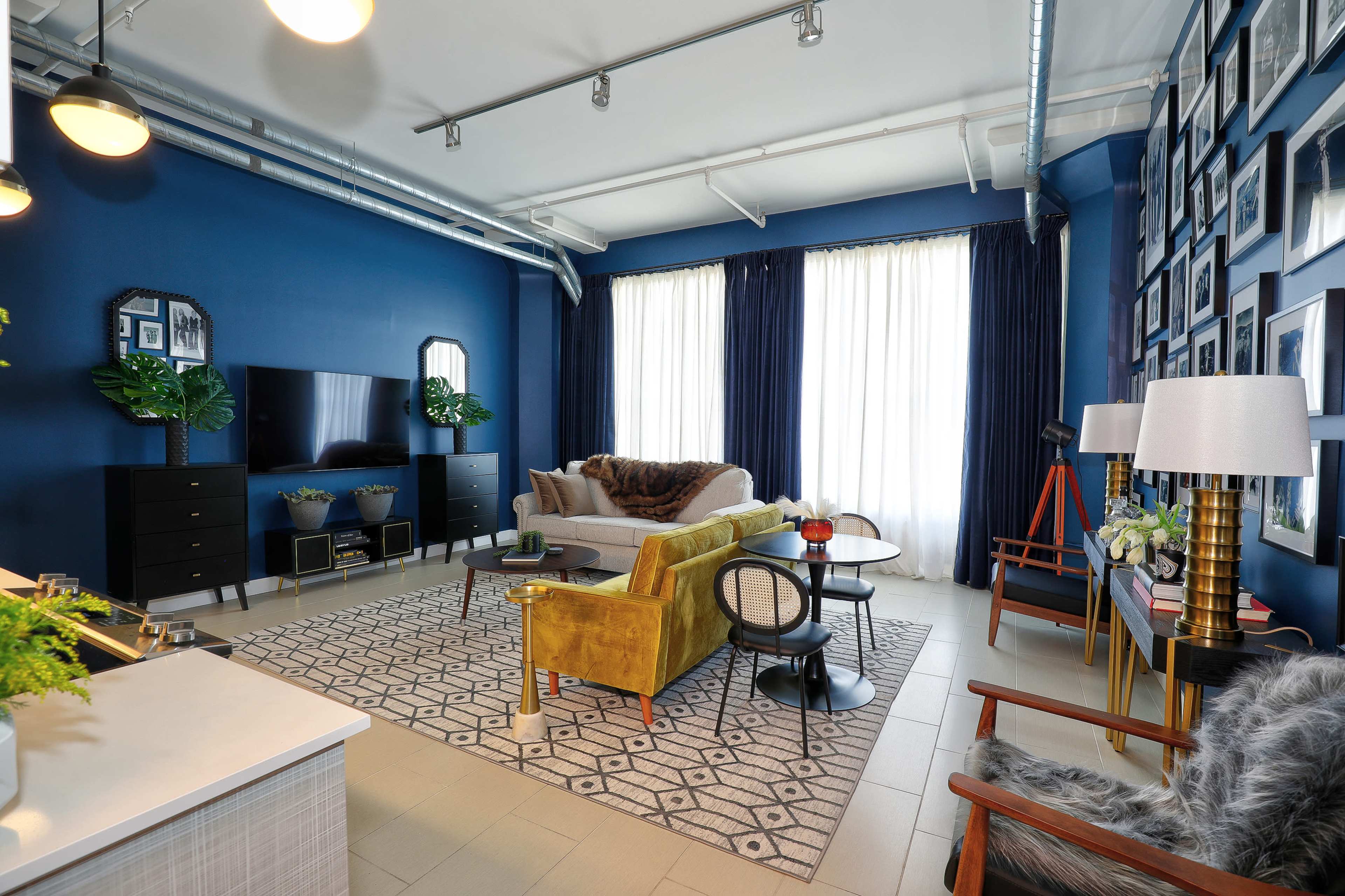 The image shows a modern room featuring a yellow sofa, a coffee table, and large windows with white curtains, surrounded by dark blue walls adorned with framed pictures.
