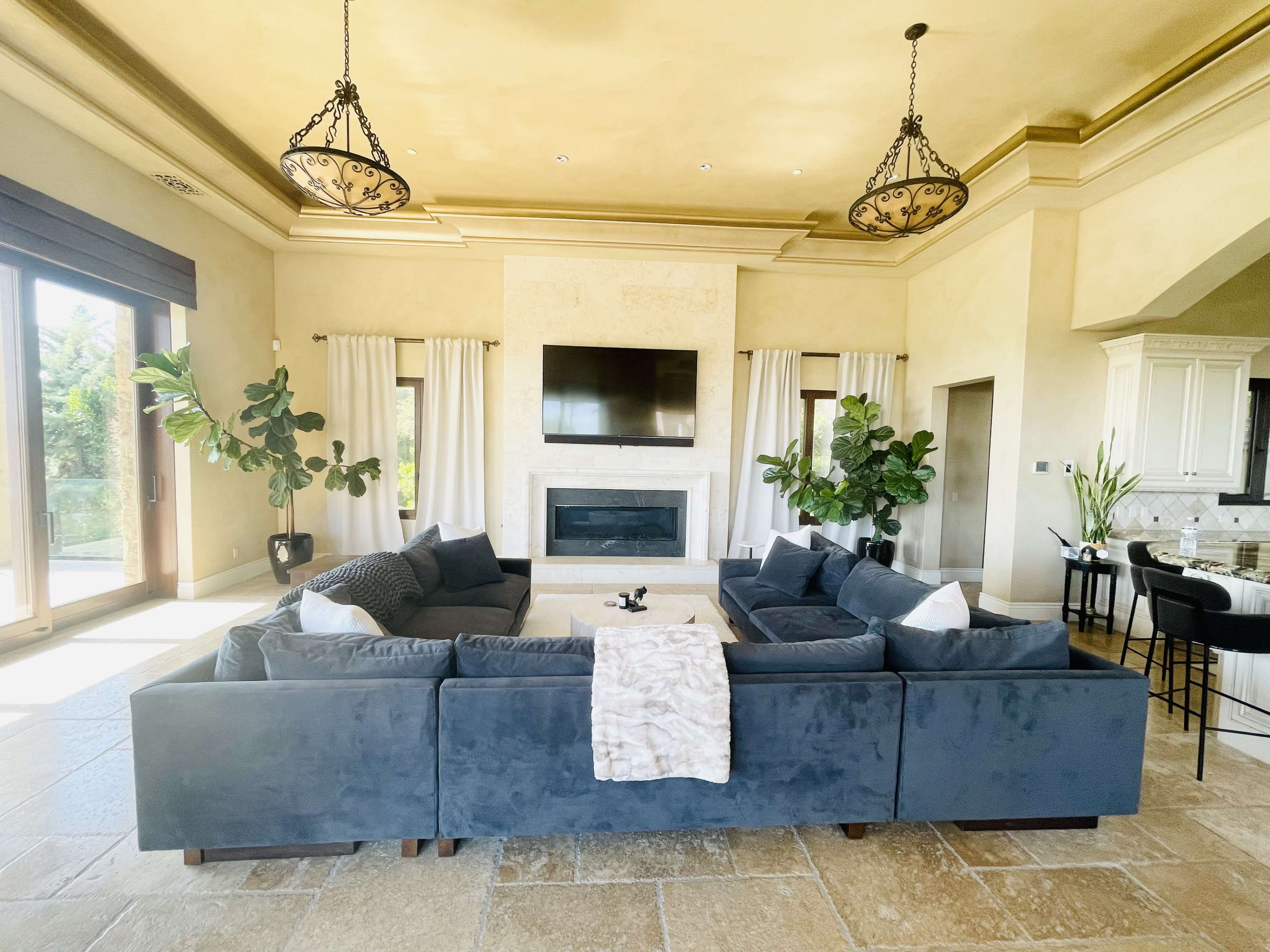 The spacious living room features a large sectional sofa arranged around a coffee table, with a fireplace and a mounted television on the wall, framed by large windows and indoor plants.