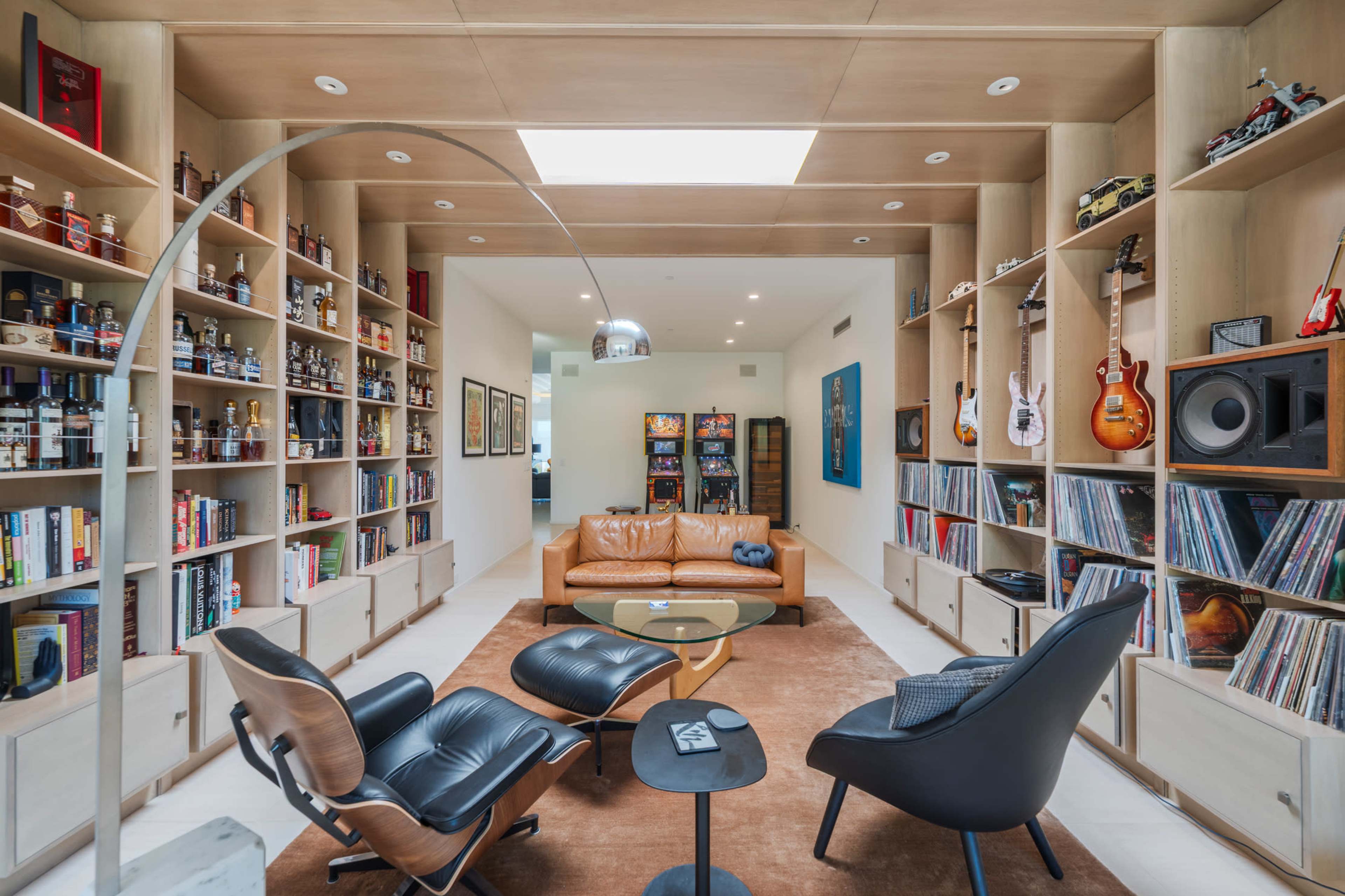 A cozy living room features a tan leather sofa, black armchairs, shelves filled with books and collectibles, and a wall of vinyl records.