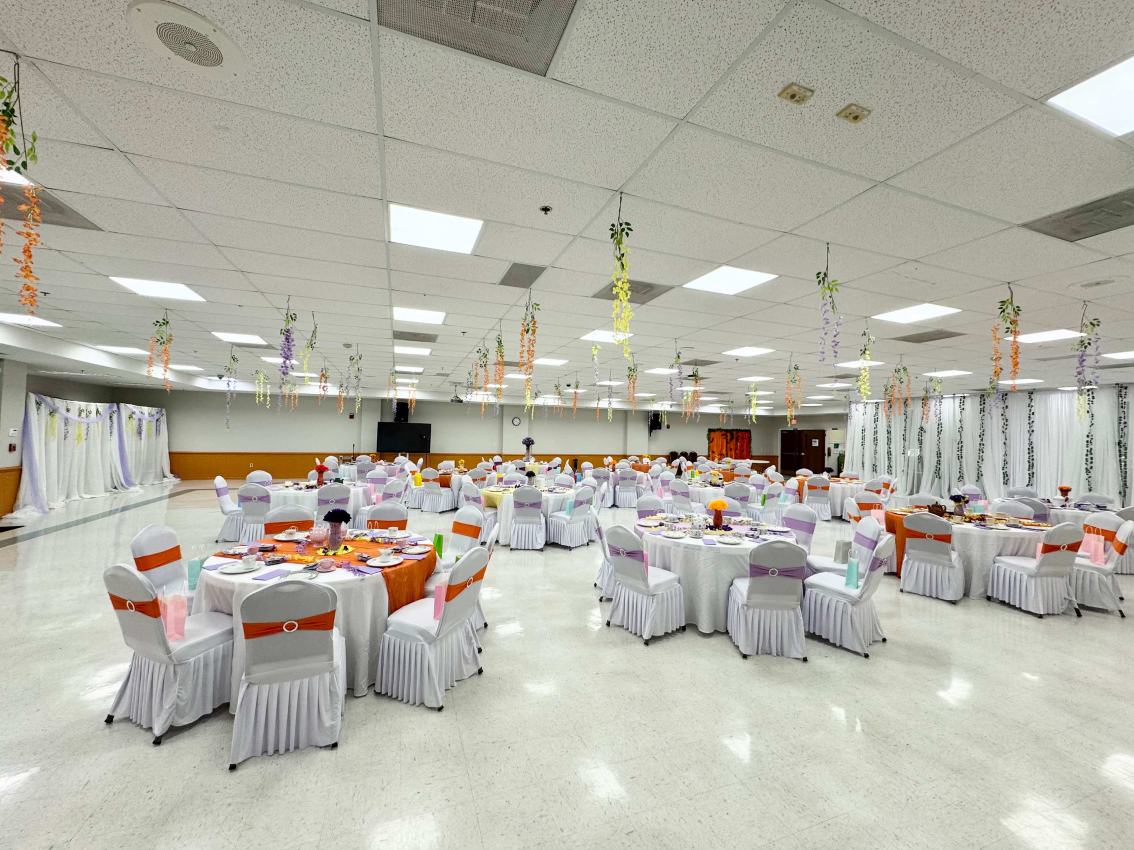 The image shows a spacious banquet hall set up with numerous round tables, each covered with colorful tablecloths and arranged in a symmetrical layout, while decorative elements hang from the ceiling.