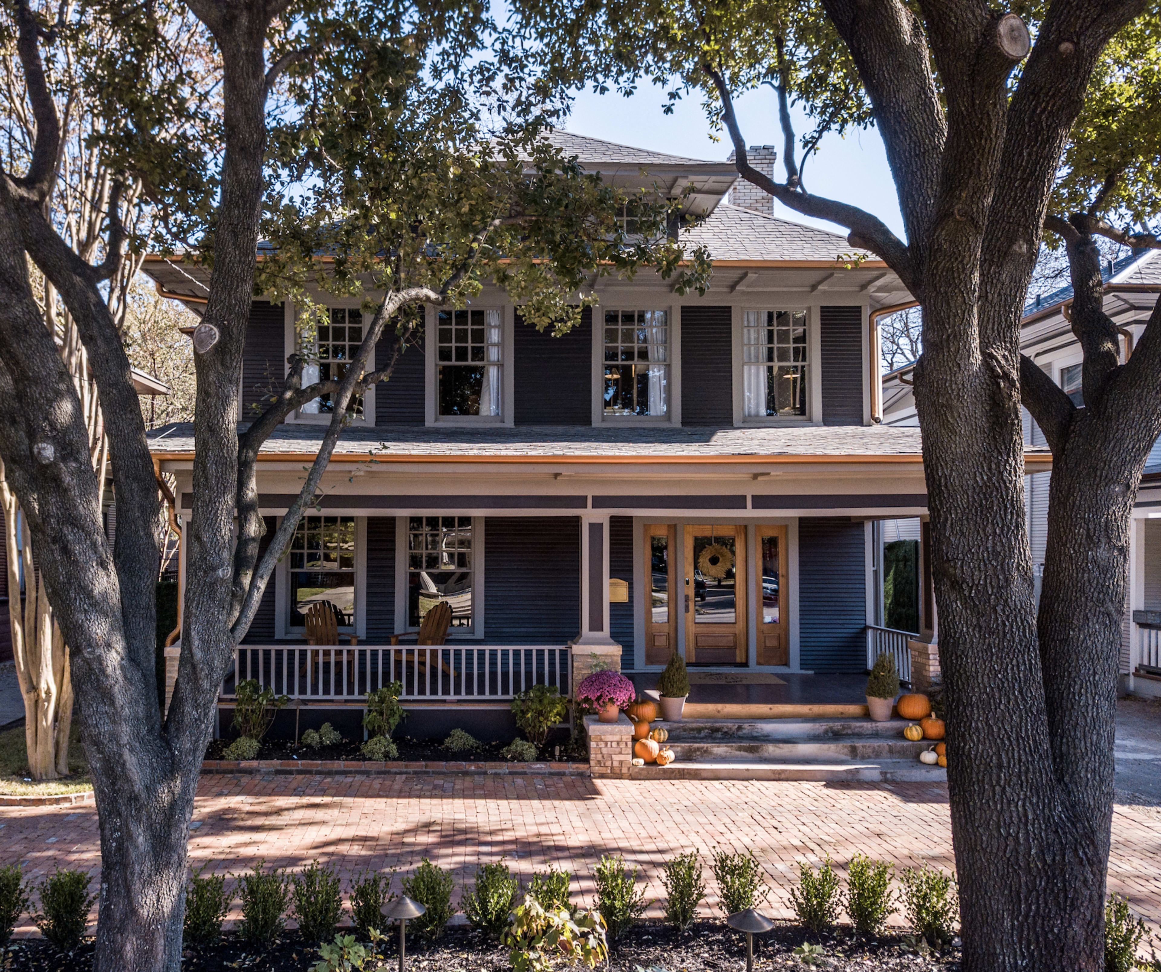 A two-story blue house with a front porch and several large windows is surrounded by trees and a landscaped garden.